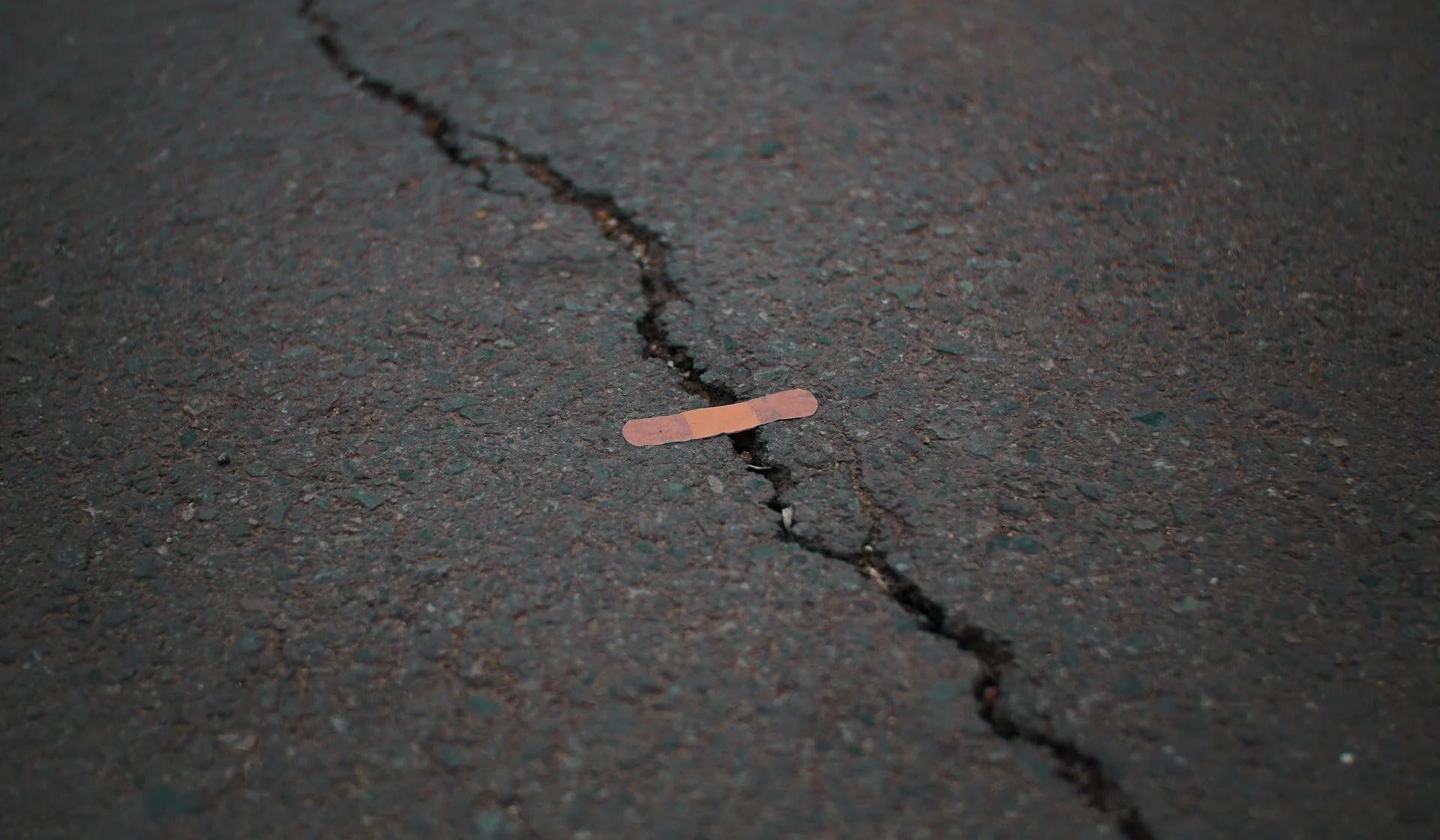 A broken asphalt street with a small, worn adhesive bandage placed in the crack.