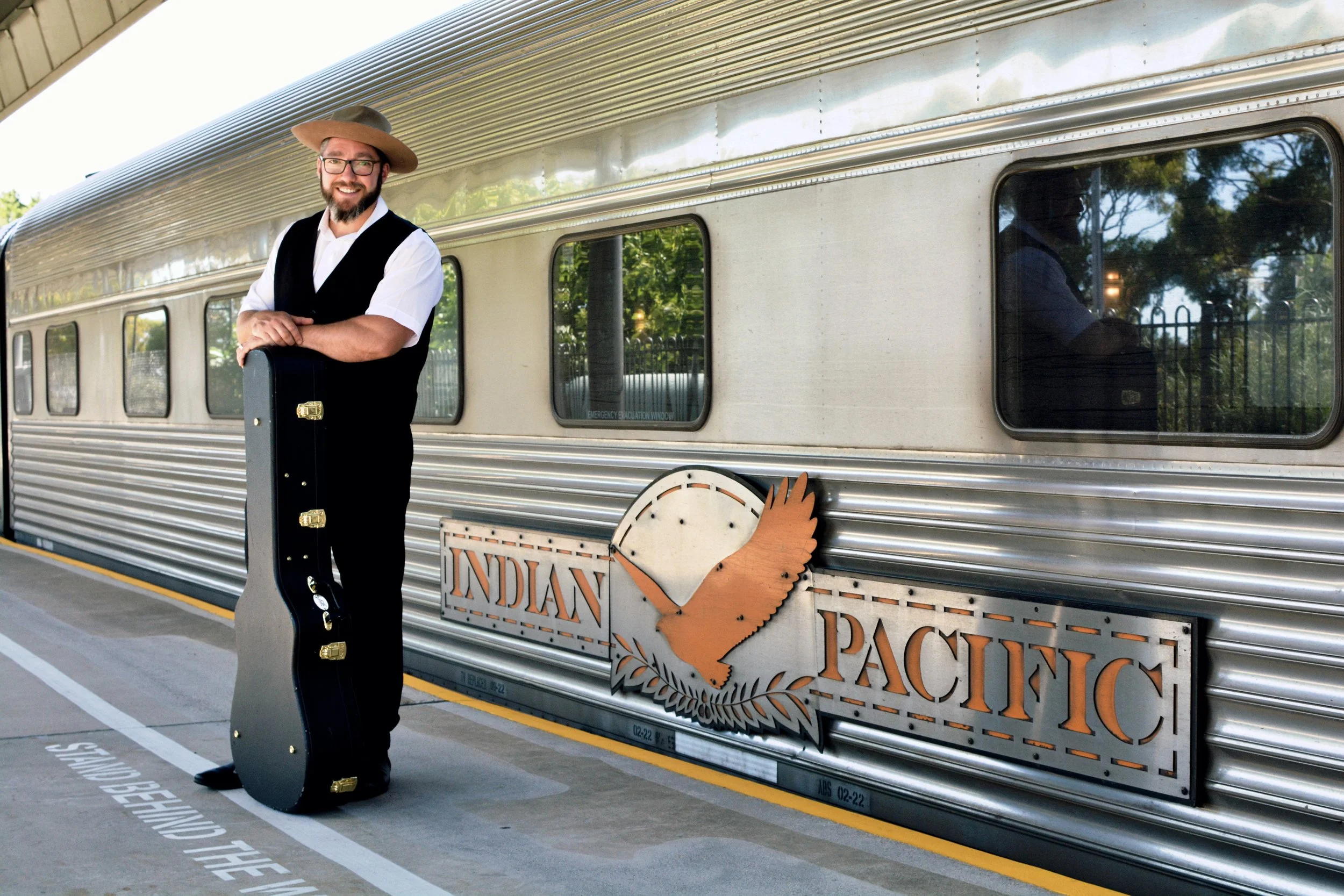 A man in a cowboy hat, white shirt, and black vest standing with a guitar case in front of an Indian Pacific train at a station platform.