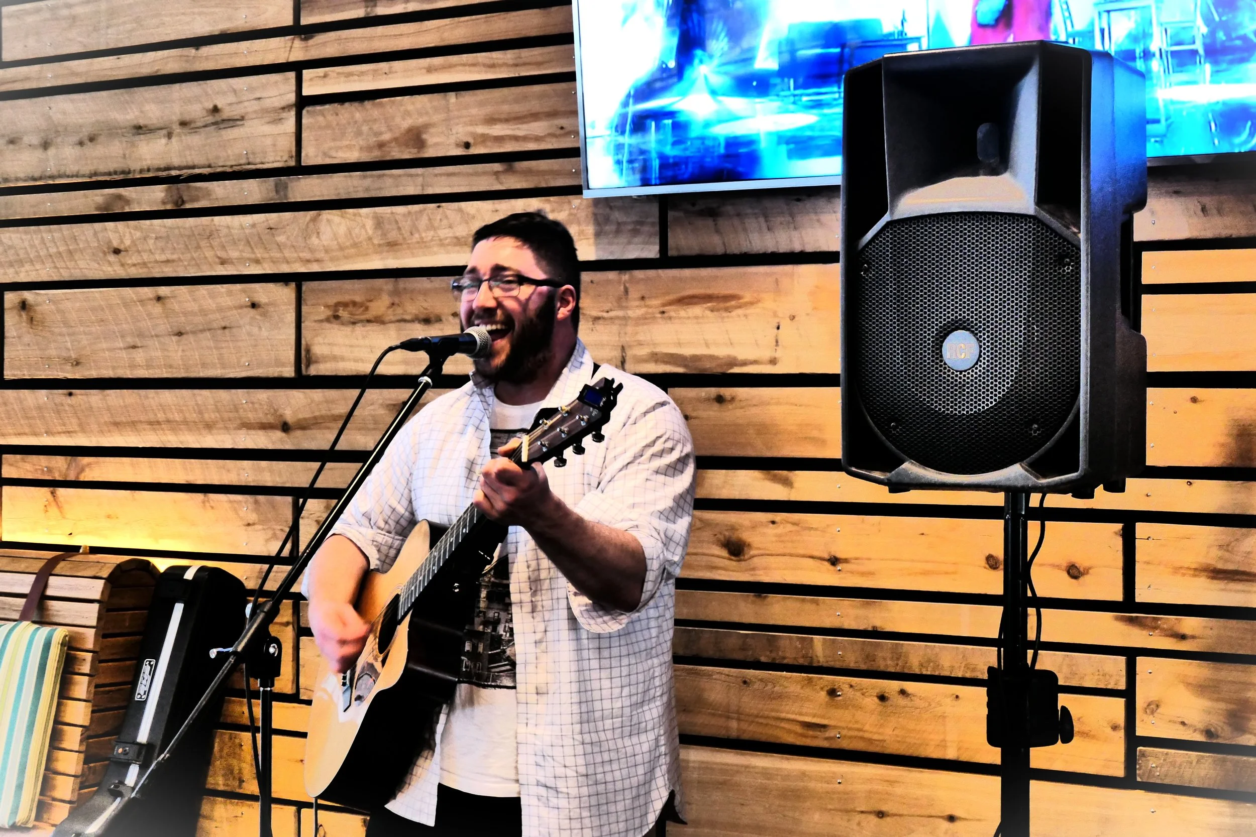 A man wearing glasses and a checkered shirt is singing into a microphone and playing an acoustic guitar. Behind him is a wooden-paneled wall, and to his right is a large black speaker. A television screen displaying vibrant blue graphics is mounted o