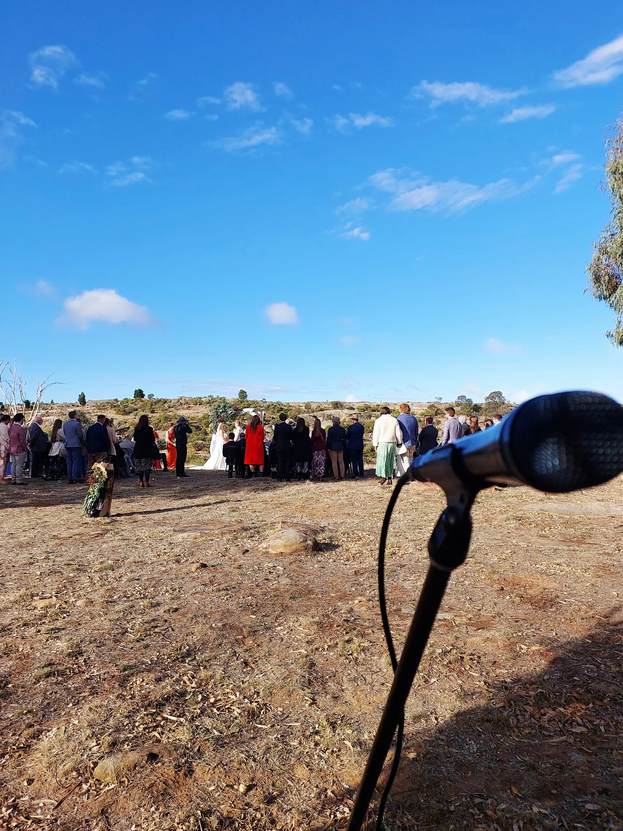 Outdoor wedding ceremony with guests standing on dirt ground under a bright blue sky with scattered clouds, microphone in foreground, and landscape in background.