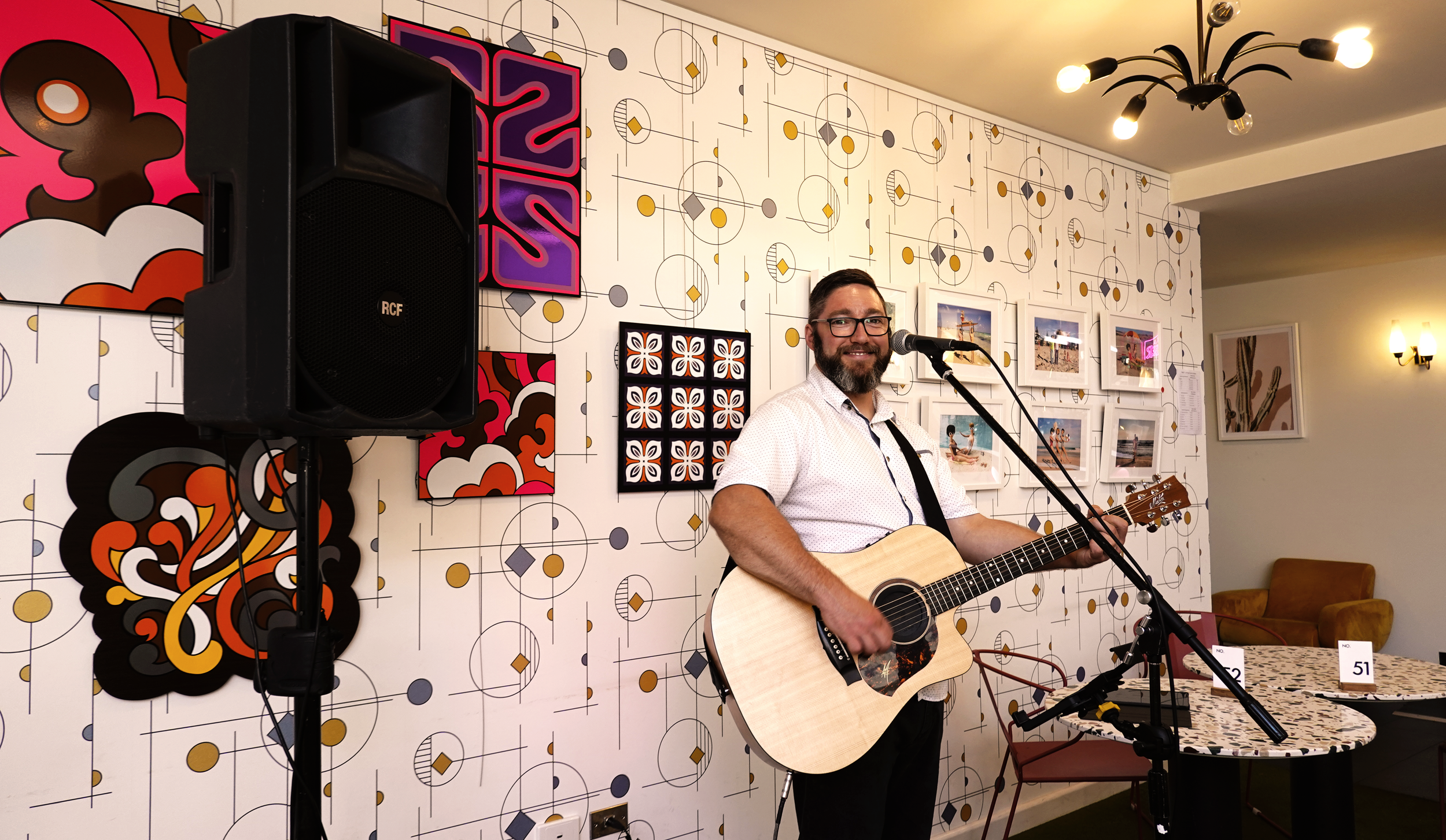 A man with glasses and a beard playing an acoustic guitar in a decorated room with framed photos and abstract art on the wall