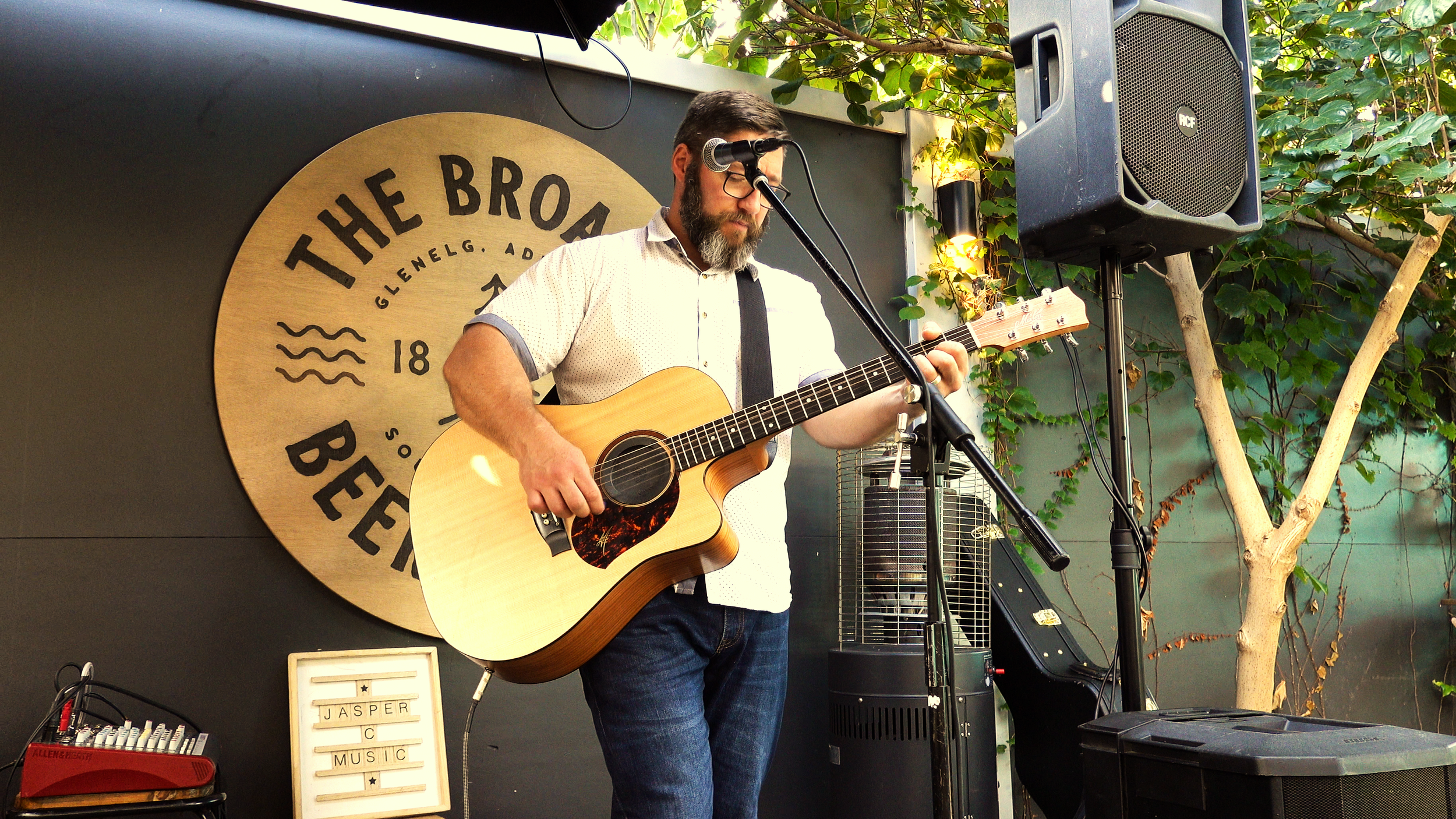 In Adelaide, Jasper C Music, wearing glasses and a beard plays an acoustic guitar on stage, singing into a microphone. The background features a circular sign that reads 'The Broad' and some greenery.