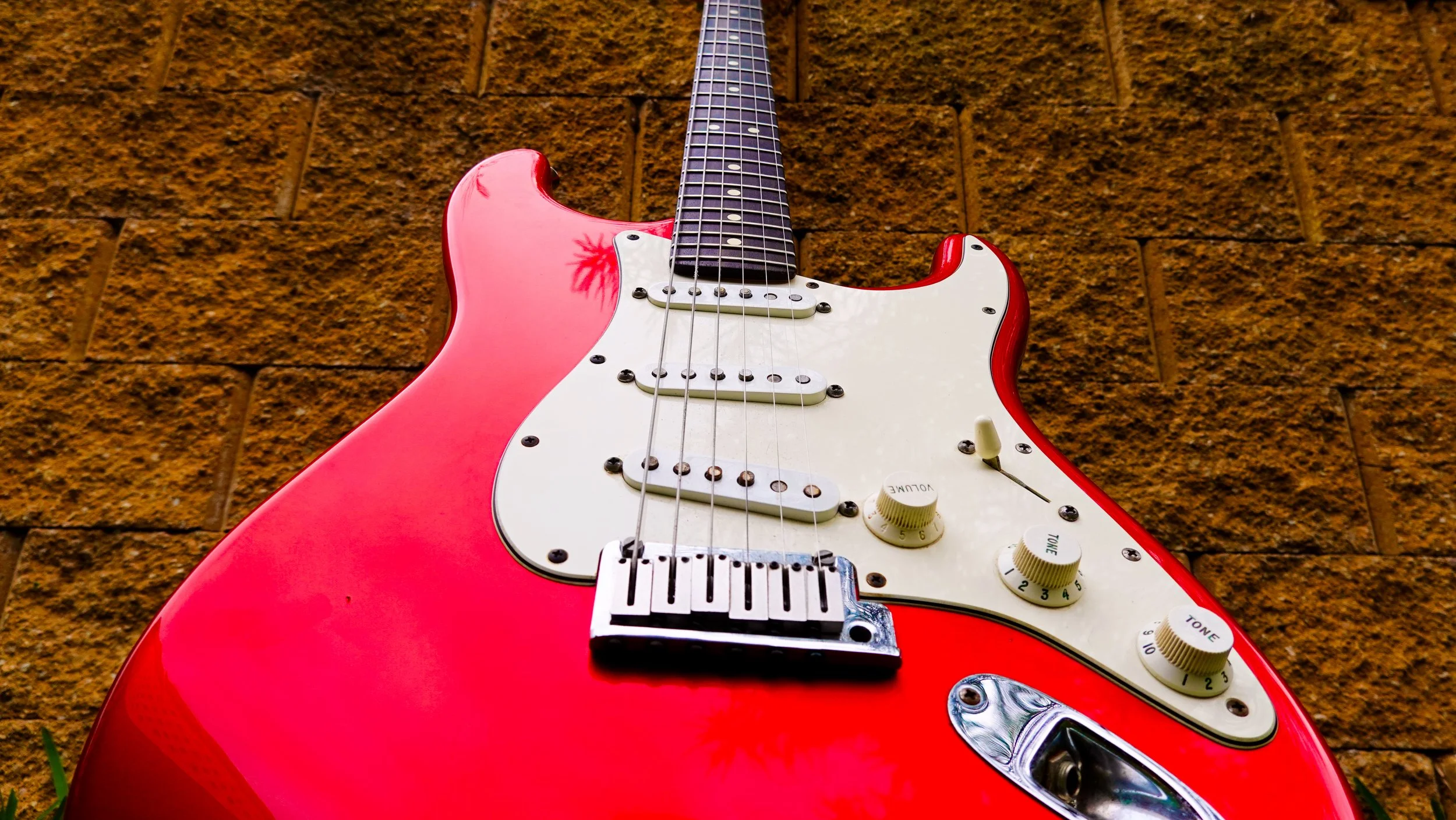 Red electric guitar with white pickguard and control knobs, mounted on an orange brick wall.