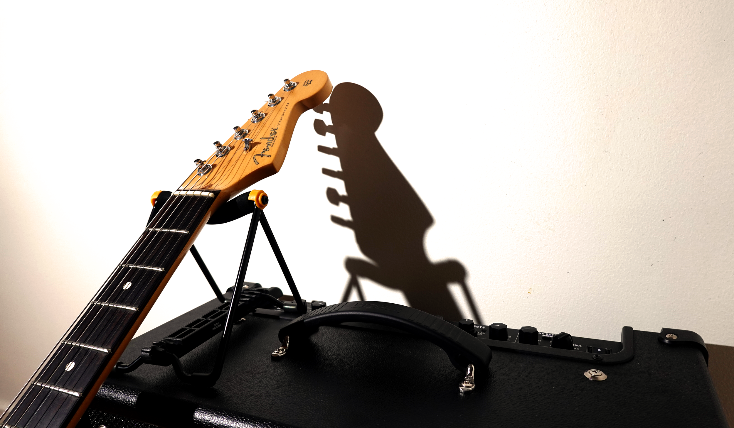 An electric guitar resting on a stand next to an amplifier, with its shadow cast on the plain white wall behind them.