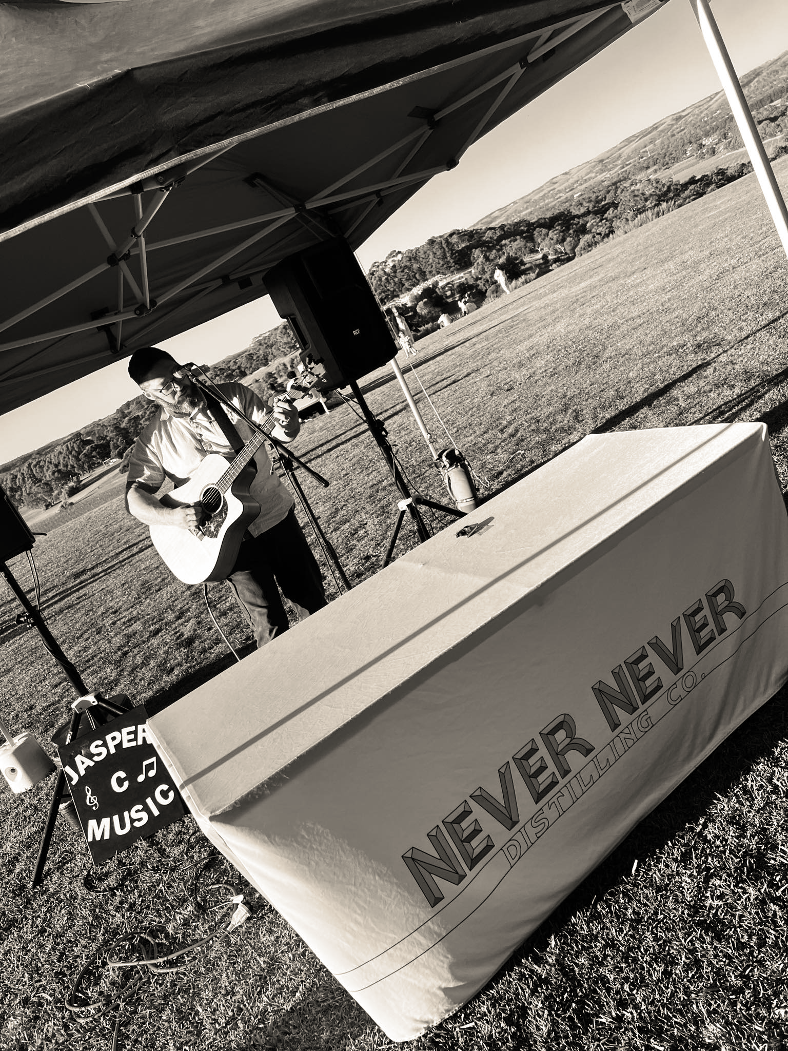In Adelaide, Jasper C Music playing guitar and singing at an outdoor event under a canopy, with a banner reading "NETHER NEVER DISTRILLING CO" and a sign that says "JASPER MUSIC," set in a grassy field with hills in the background.