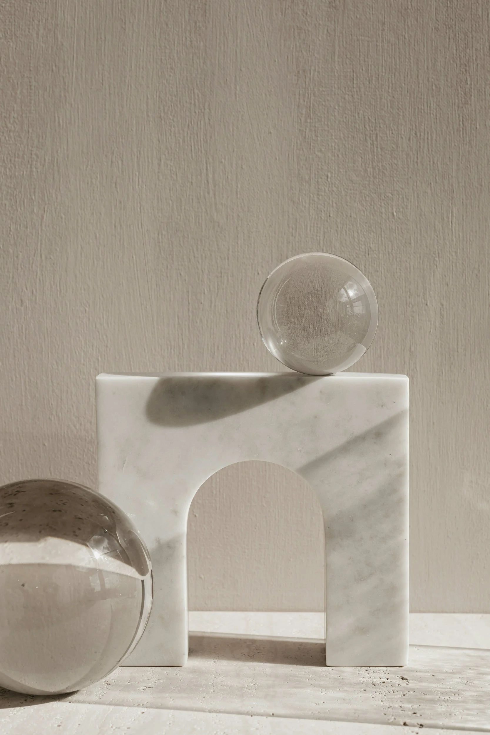 Minimalist composition of two glass spheres and a marble-like arch structure on a neutral surface, with a plain beige background.