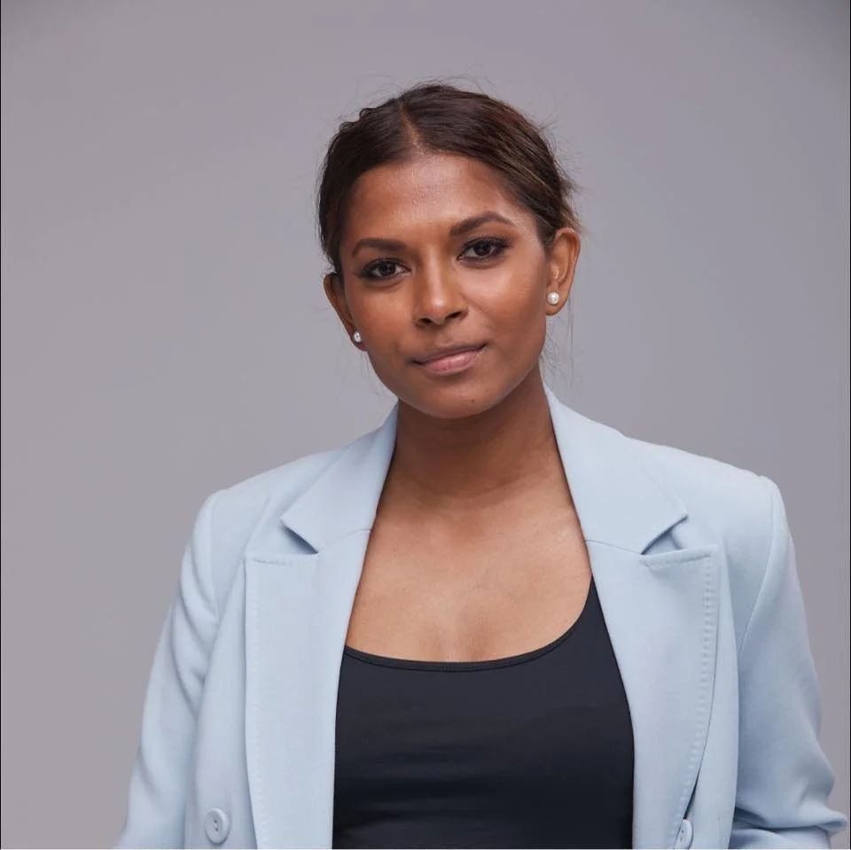 A woman with medium brown skin, wearing a light gray blazer and a black top, posing against a plain gray background.