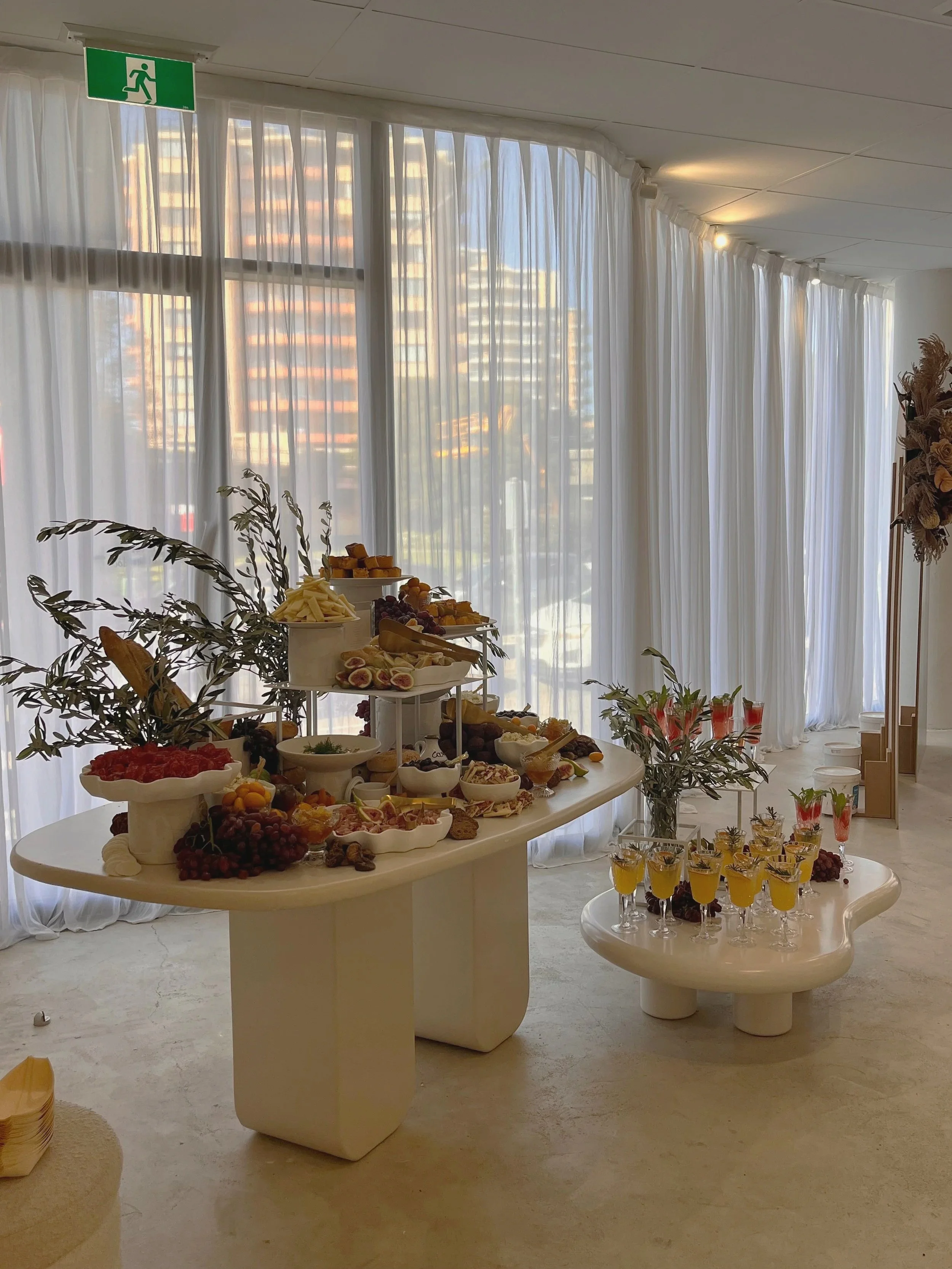 Indoor buffet with an assortment of snacks and drinks, decorated with plants and white curtains, with city buildings visible outside the window.