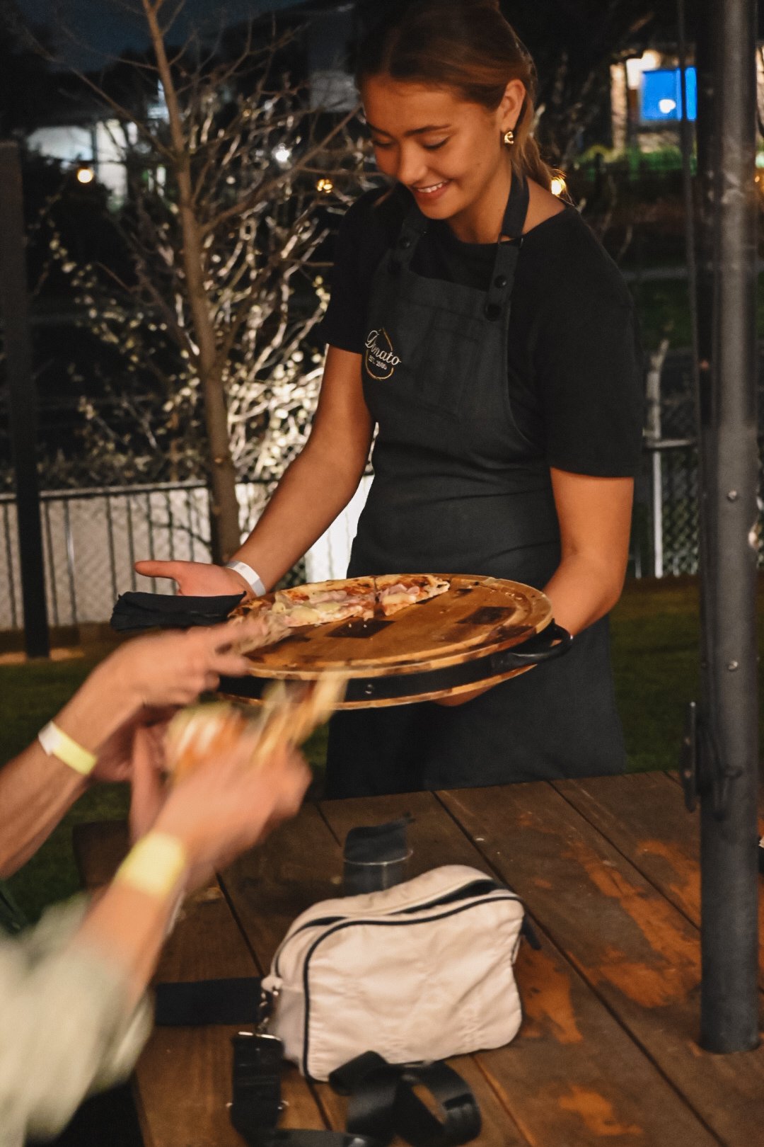 A woman in a black shirt and apron serving pizza on a wooden pizza peel to a person reaching for a slice, outdoors at night.