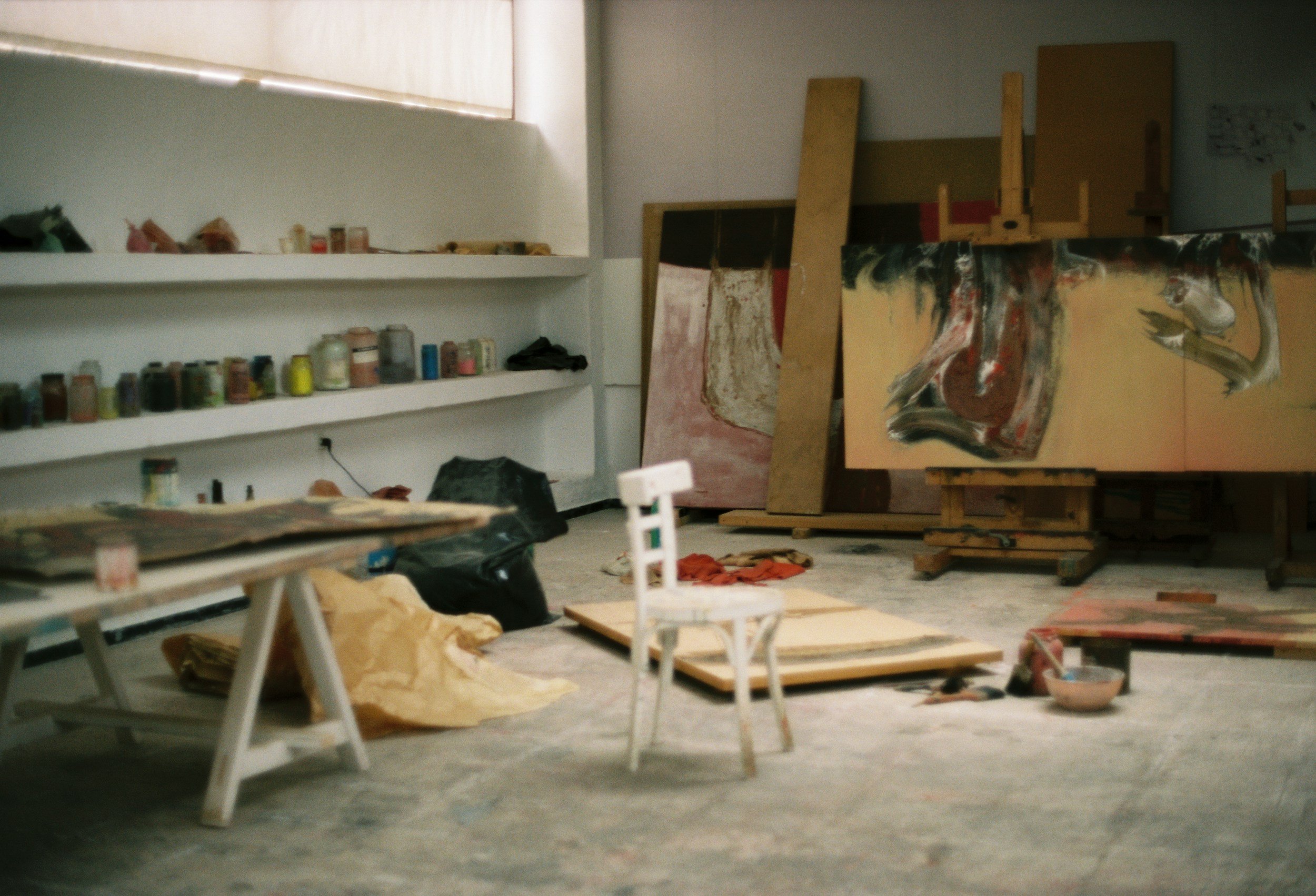 An art studio with paintings, art supplies on shelves, and tables, in a somewhat cluttered space. Studio Lee Jae