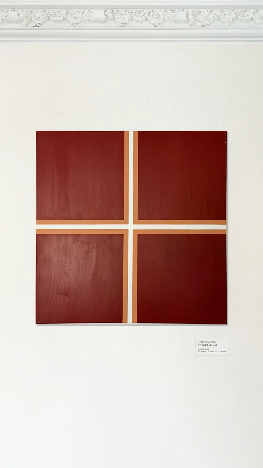 A square piece of artwork with four red panels framed with beige lines, mounted on a white wall with ornate molding at the top.