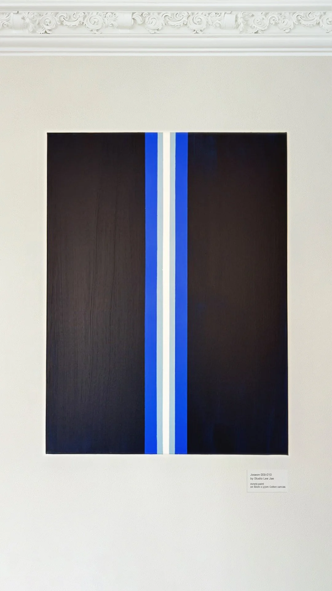 Abstract painting with a vertical black rectangle, bordered by white, with a thin blue line on each side, displayed on a white wall with ornate crown molding.