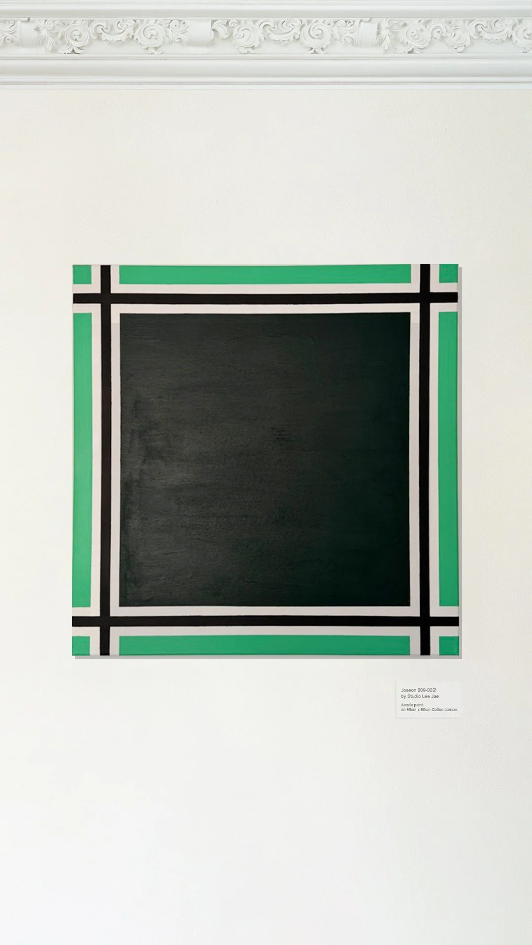A black square with a decorative border of green, black, and white in a geometric pattern, mounted on a white wall with ornate crown molding above.