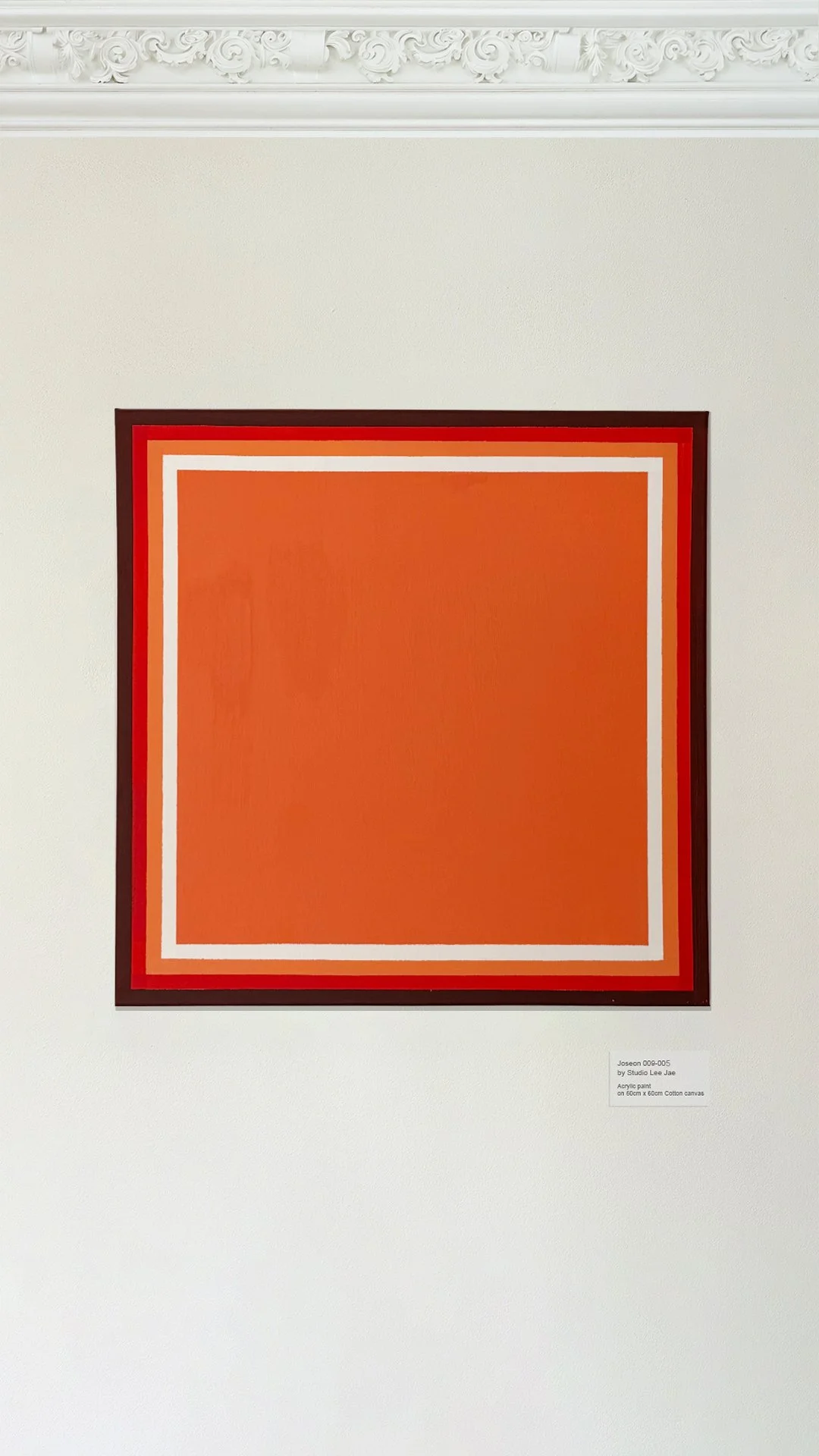 A framed geometric abstract painting with a large orange square centered, surrounded by white and red borders, hanging on a white gallery wall with decorative molding.