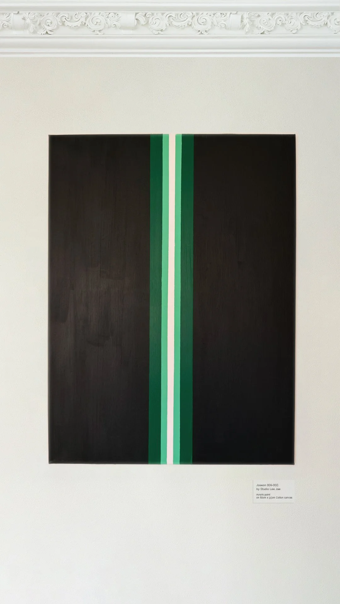 A rectangular black canvas with vertical green, white, and light green stripes in the center, displayed on a white wall with decorative molding at the top.
