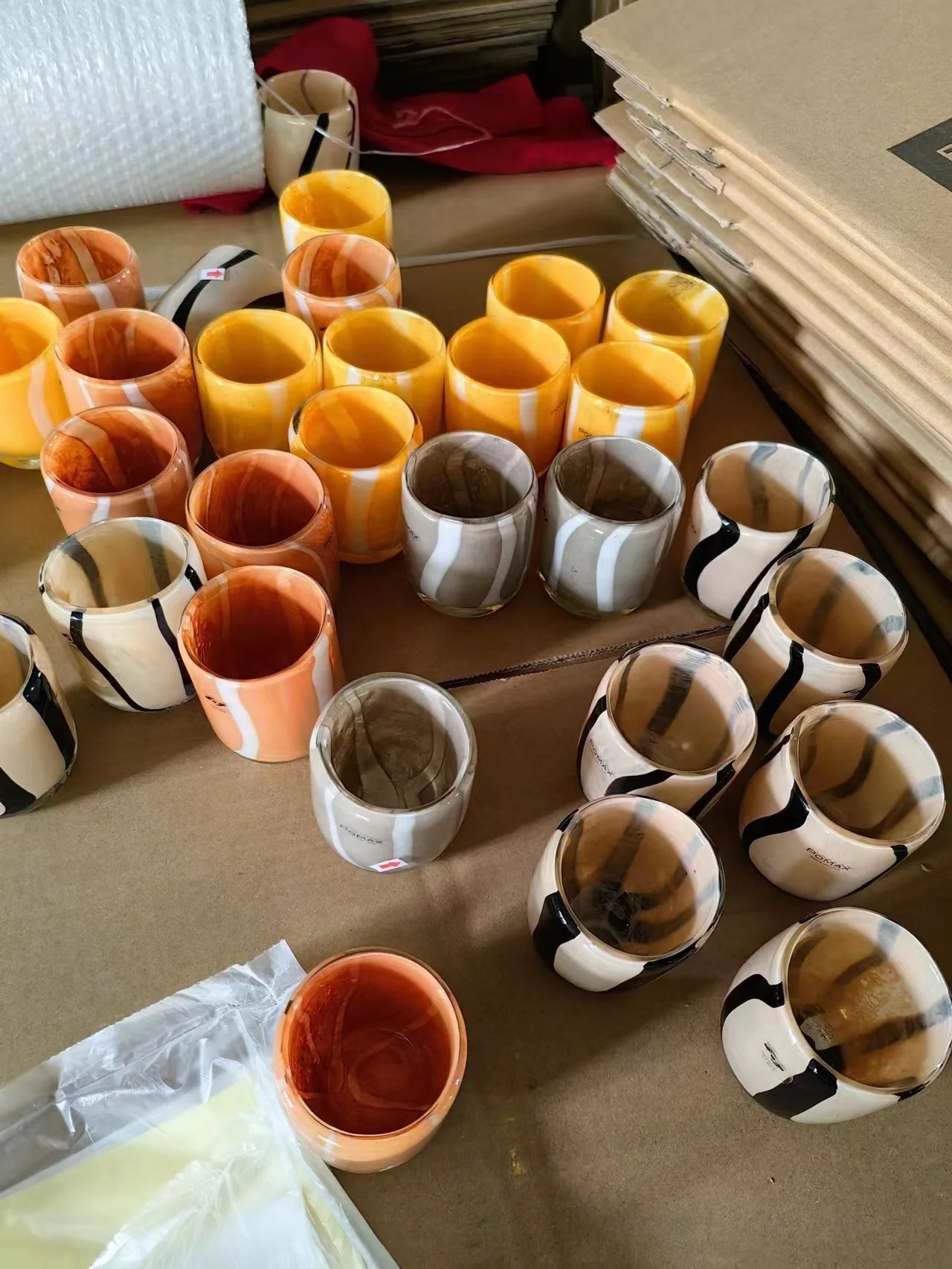 An assortment of small, colorful ceramic cups in shades of yellow, orange, white, and black on a table, with some stacked plates and supplies in the background.