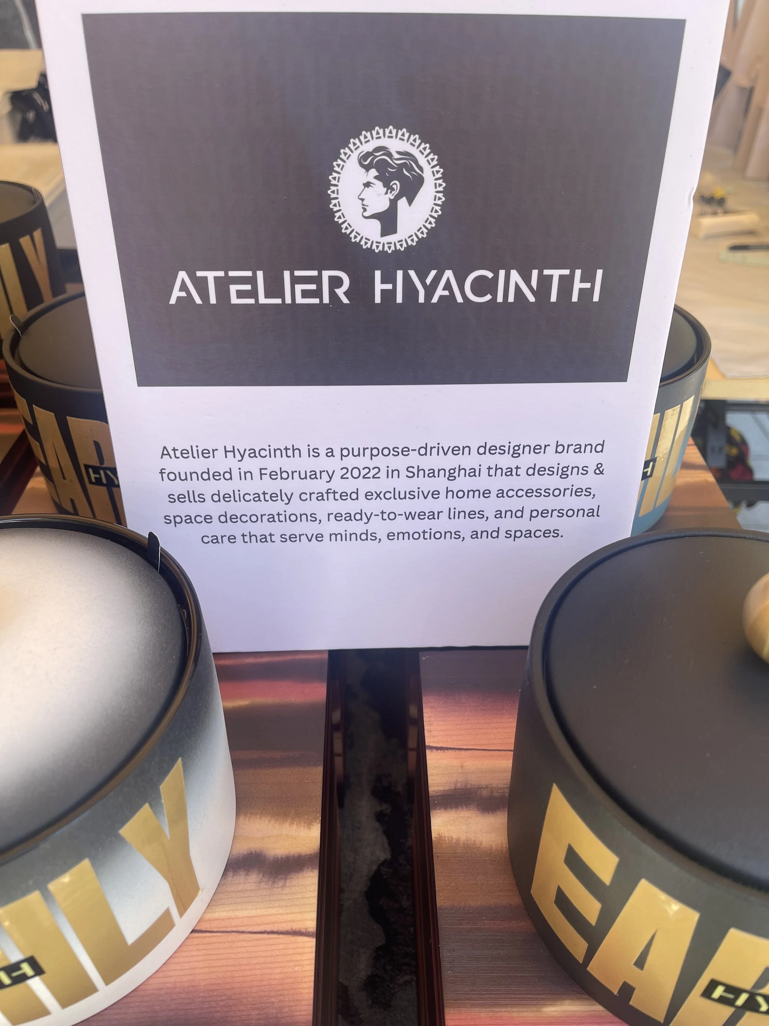 Sign for Atelier Hyacinth with description about the brand, located on a wooden table with black and yellow containers nearby.