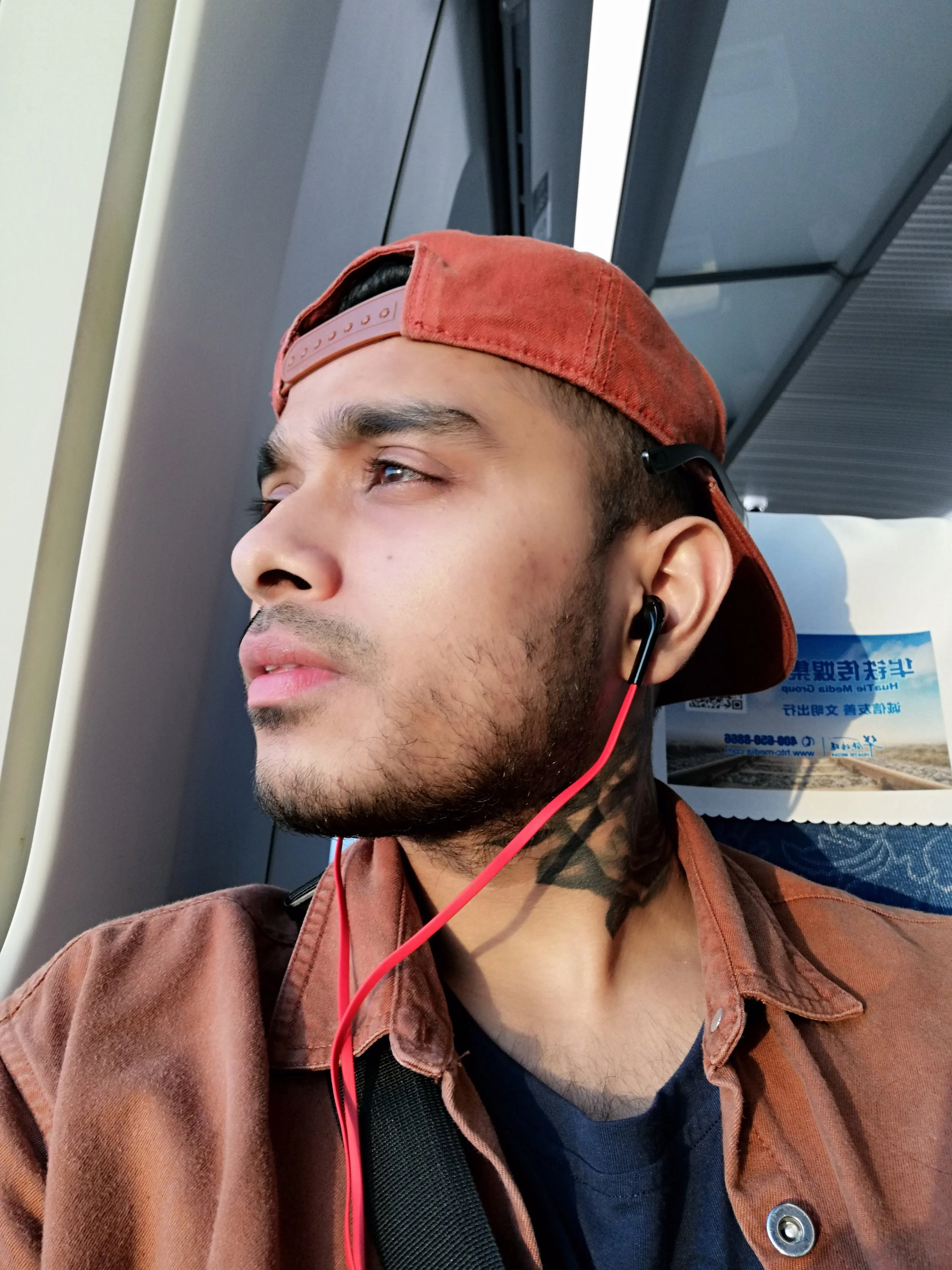 A young man wearing a red baseball cap backward and black earbuds with red cords, looking out the window on a train.