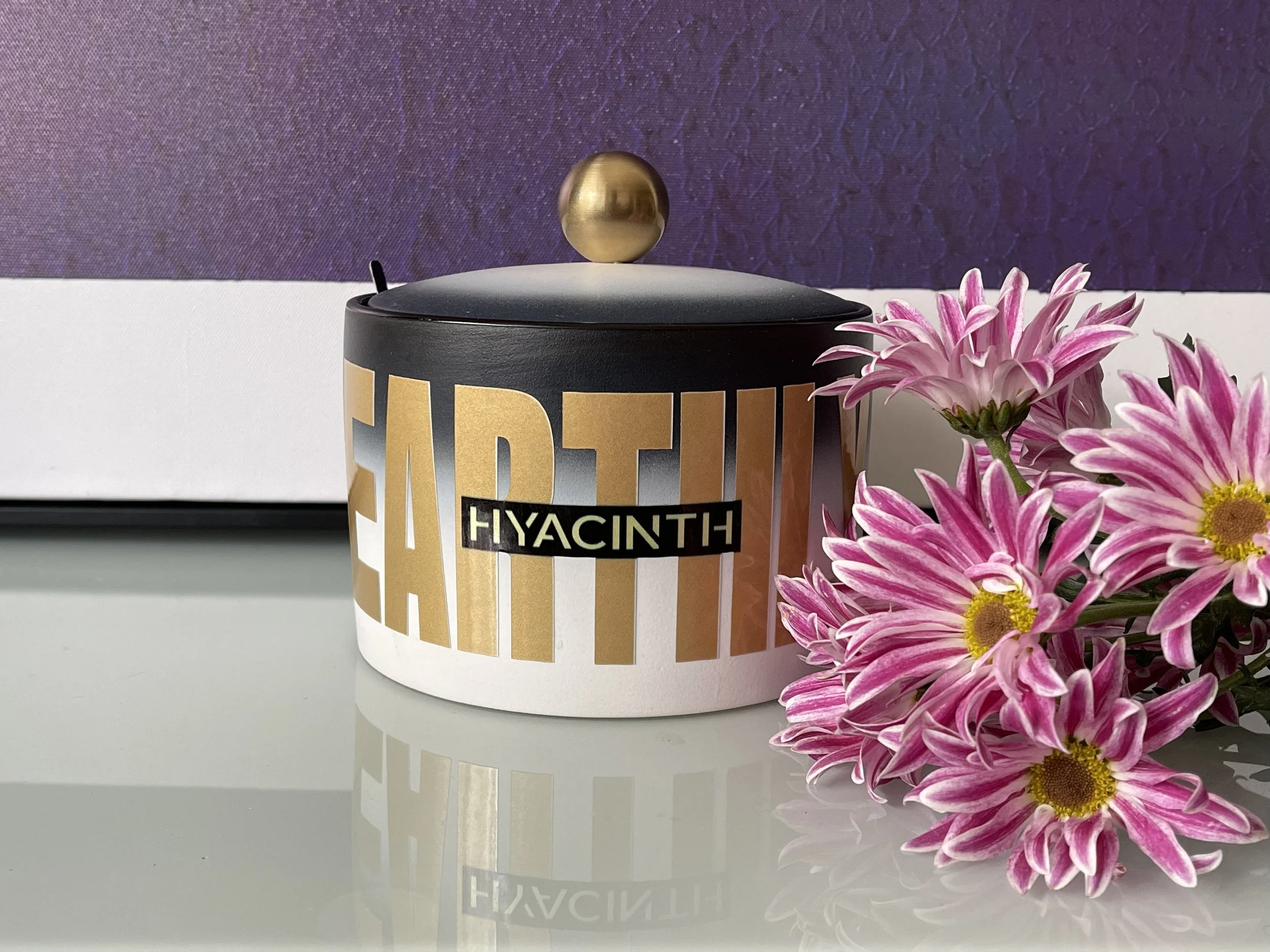 A cylindrical container with a black lid, gold and white labels, and a gold knob on top, placed next to pink flowers with yellow centers, on a reflective surface with a purple and white background.