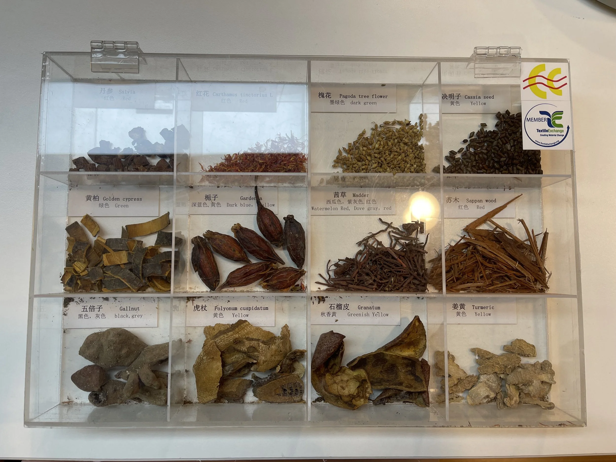 A display case with various labeled dried herbs and plants, including salvia, carthamustinctorius, pagoda tree flower, cassia seed, golden cypress, gardenia, madder, sappan wood, gallnut, polynum cuspidatum, granatum, and turmeric.