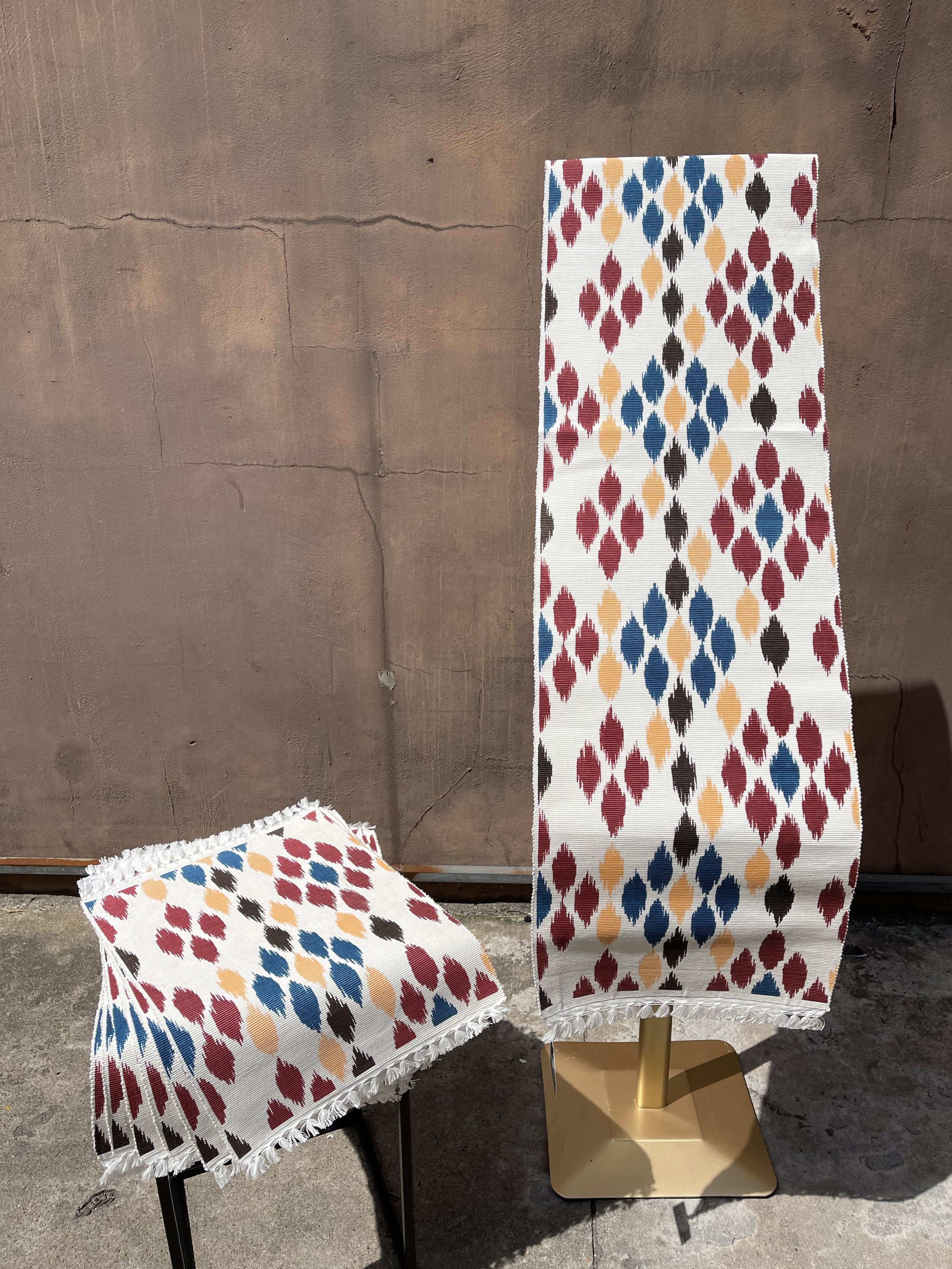 Two textiles with a colorful diamond pattern, one draped over a tall stand and the other folded on a small stool, against a brown wall.