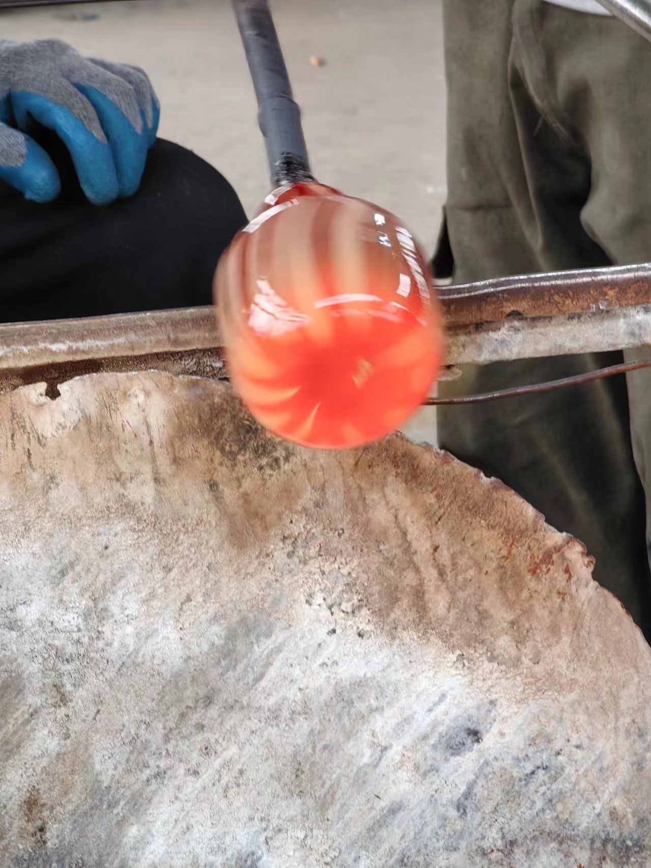 Close-up of a glassblowing process where molten glass is being shaped. The glass is glowing orange-red, indicating high heat, and is being worked on with tools. The background shows part of a work table or furnace and a person's clothing.