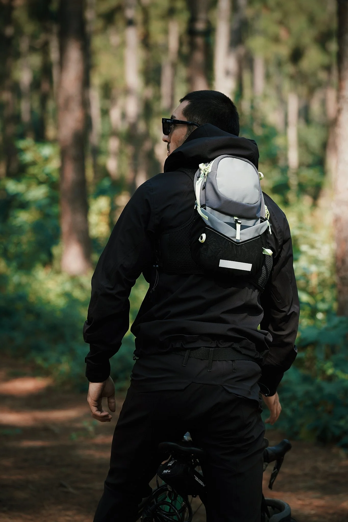 A person wearing black clothes, sunglasses, and a backpack riding a bicycle through a forest.