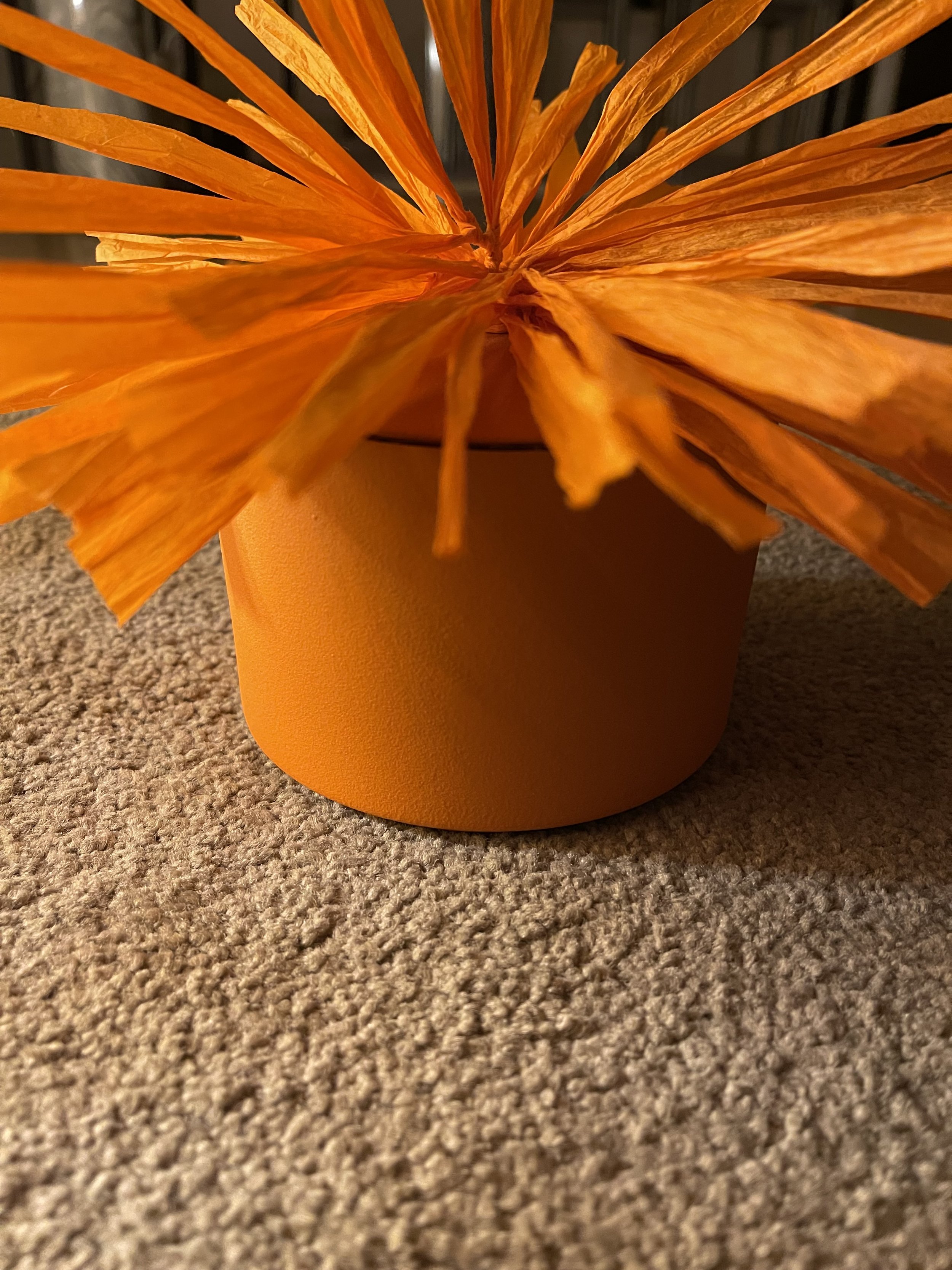 Close-up of an orange paper flower in a yellow vase on a beige carpet.