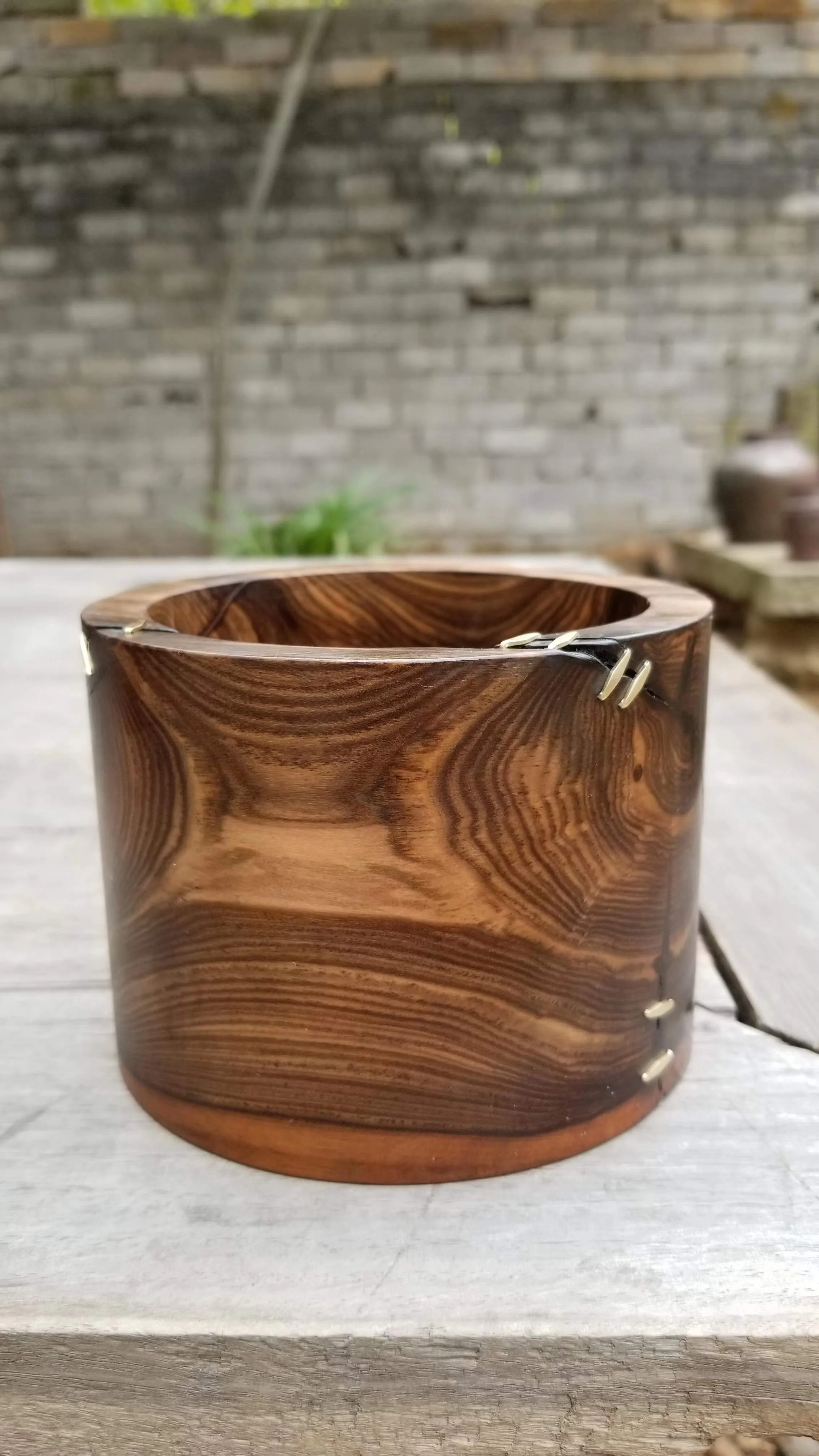 A polished wooden bracelet with metal clasp resting on a wooden surface outdoors, in front of a brick wall with some plants and pottery in the background.