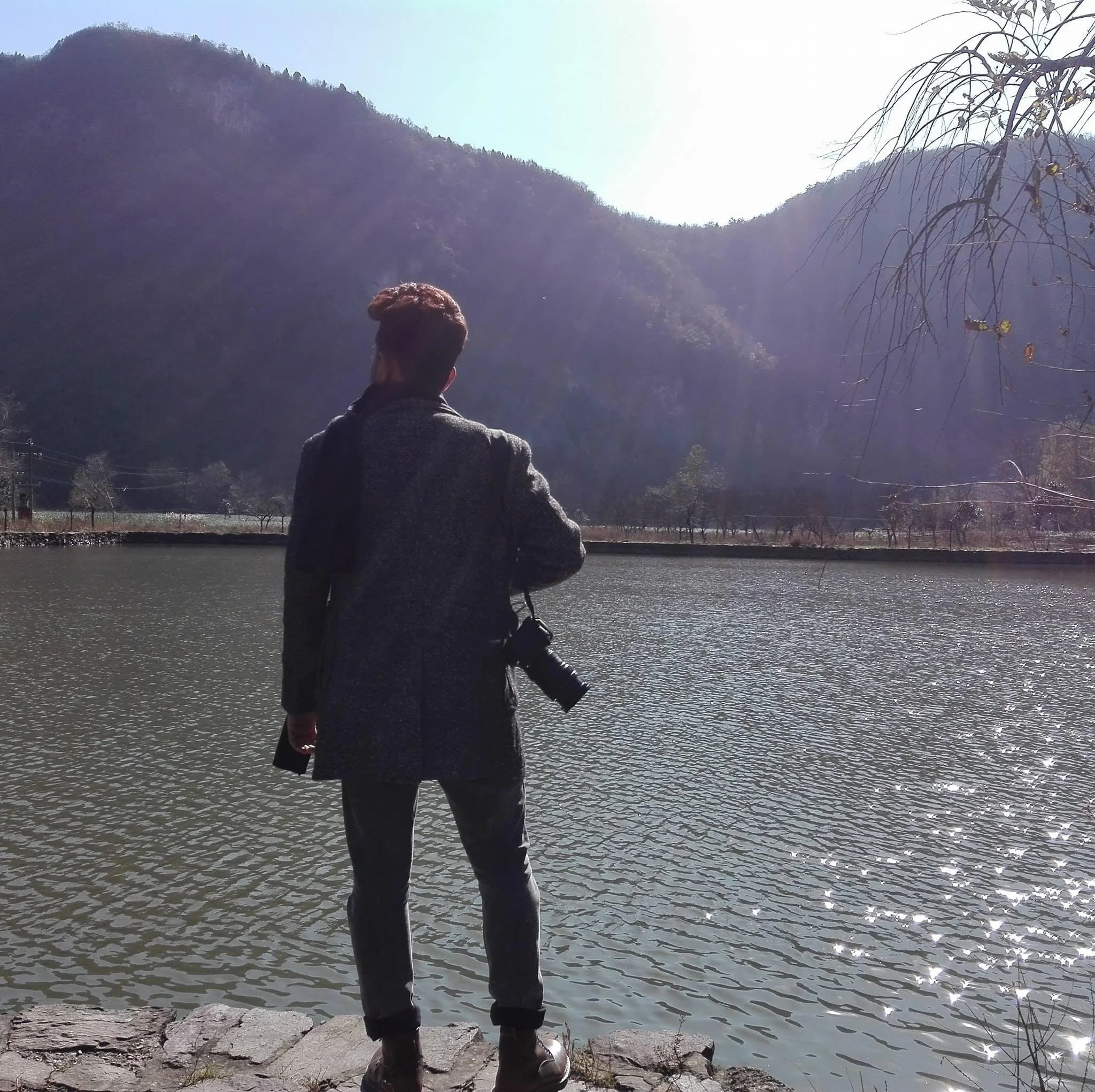 Person standing on a stone platform near a body of water with mountains and trees in the background, holding a camera, facing away from the camera.