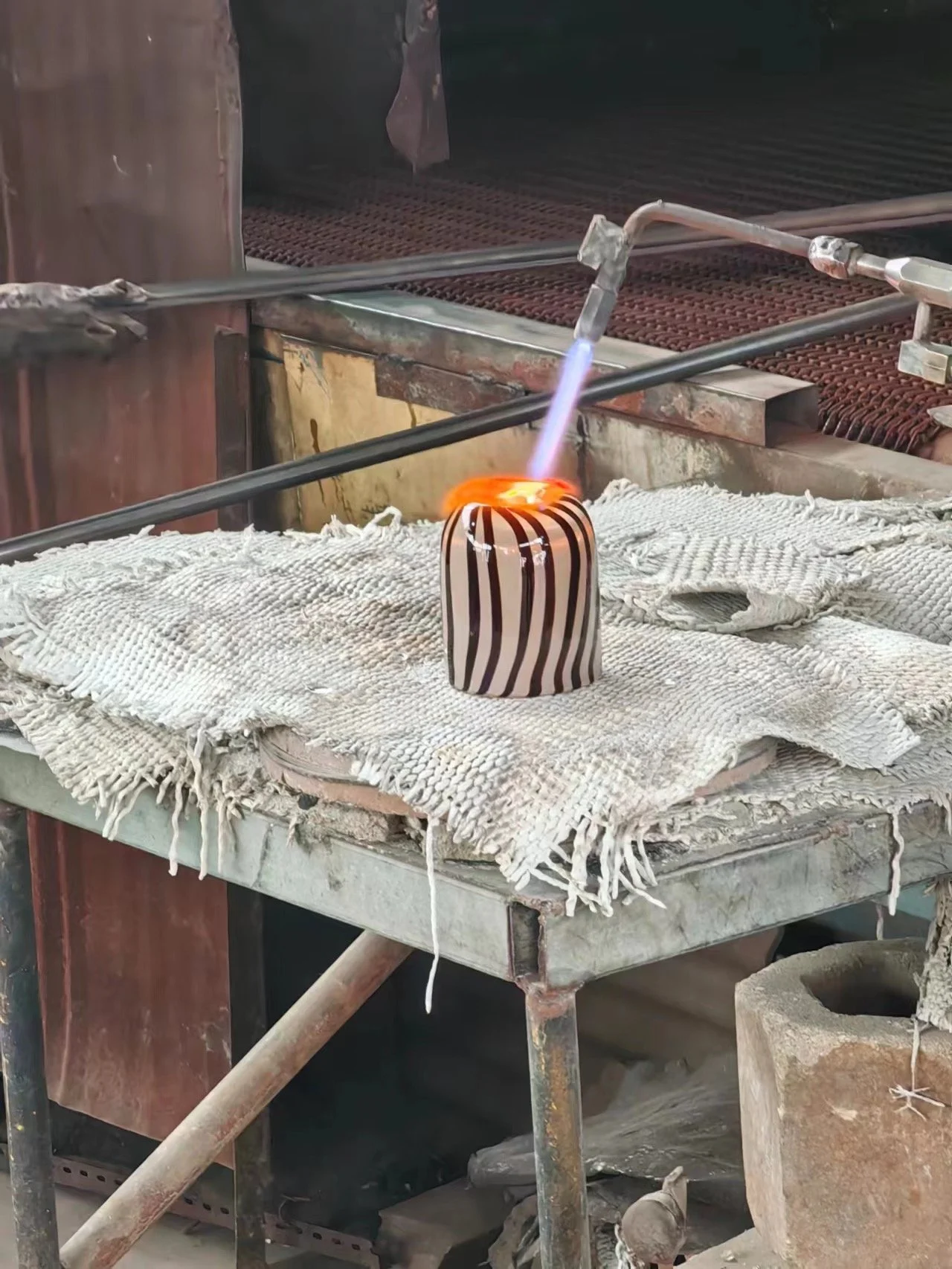 A glassblowing workshop shows a striped glass vessel on a cloth-covered worktable, with a heated blowtorch aimed at it.