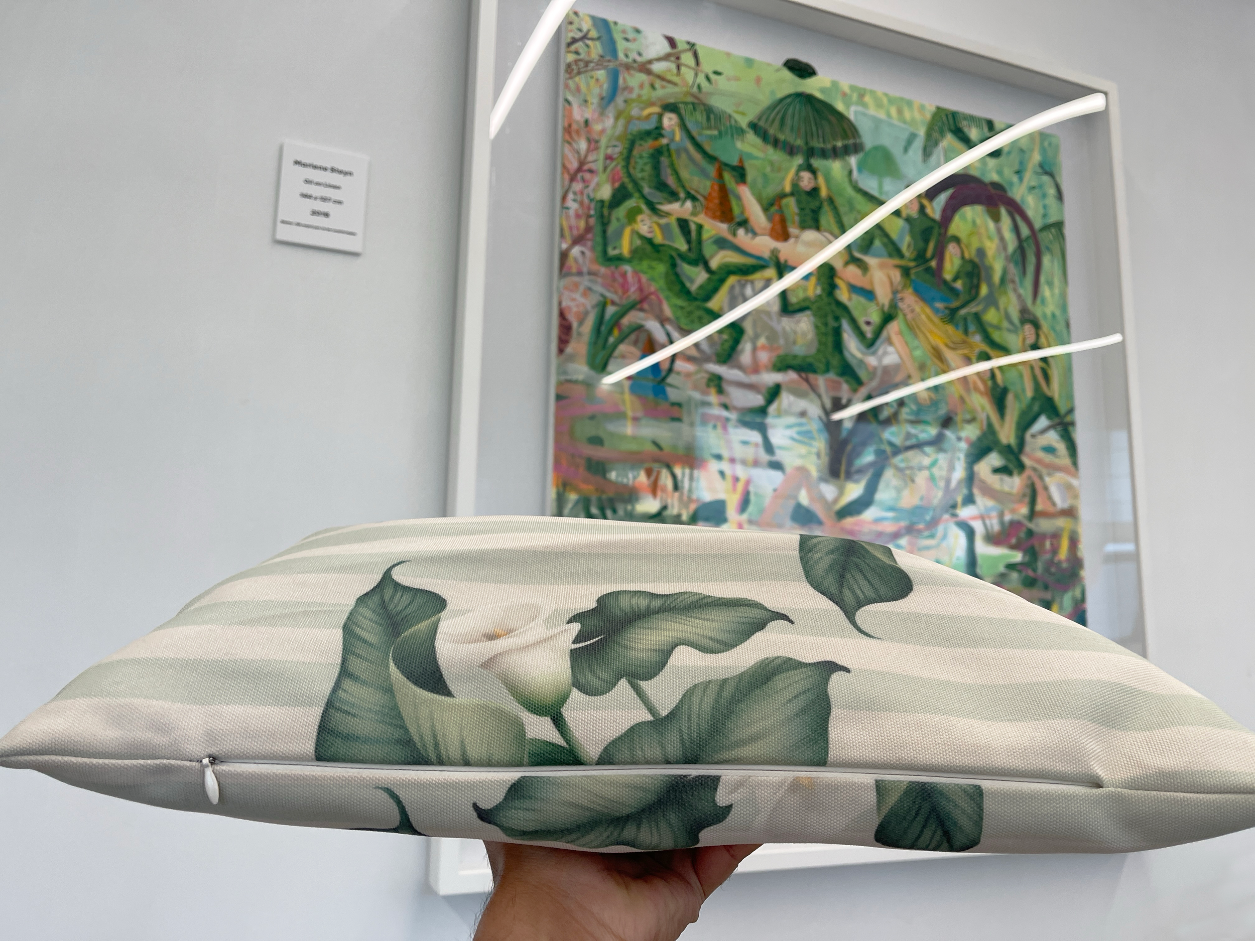 A hand holding a rectangular cushion with green leafy pattern in front of a framed colorful painting with various figures and green foliage in an art gallery.