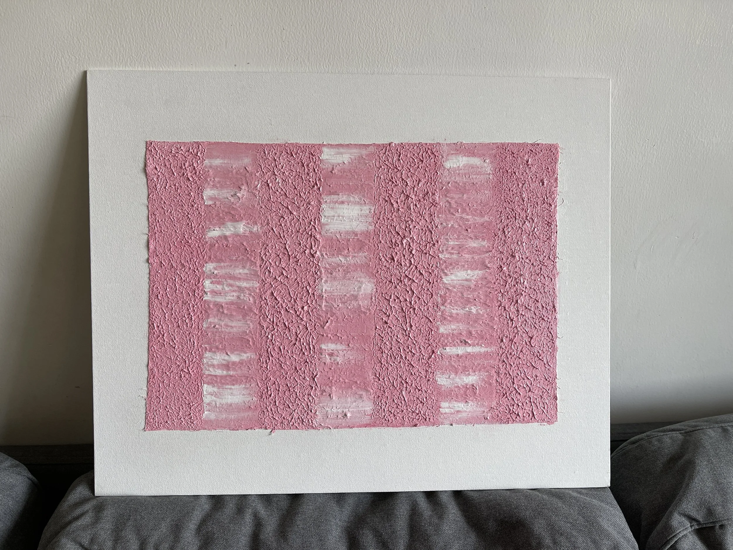 Abstract textured artwork with pink and white colors on a canvas, leaning against a wall.