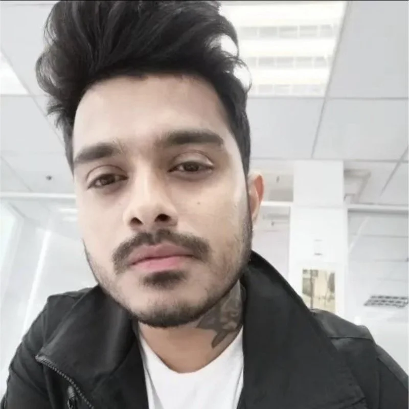A young man with dark hair and a beard in an indoor setting, wearing a black jacket and white shirt, looking at the camera.