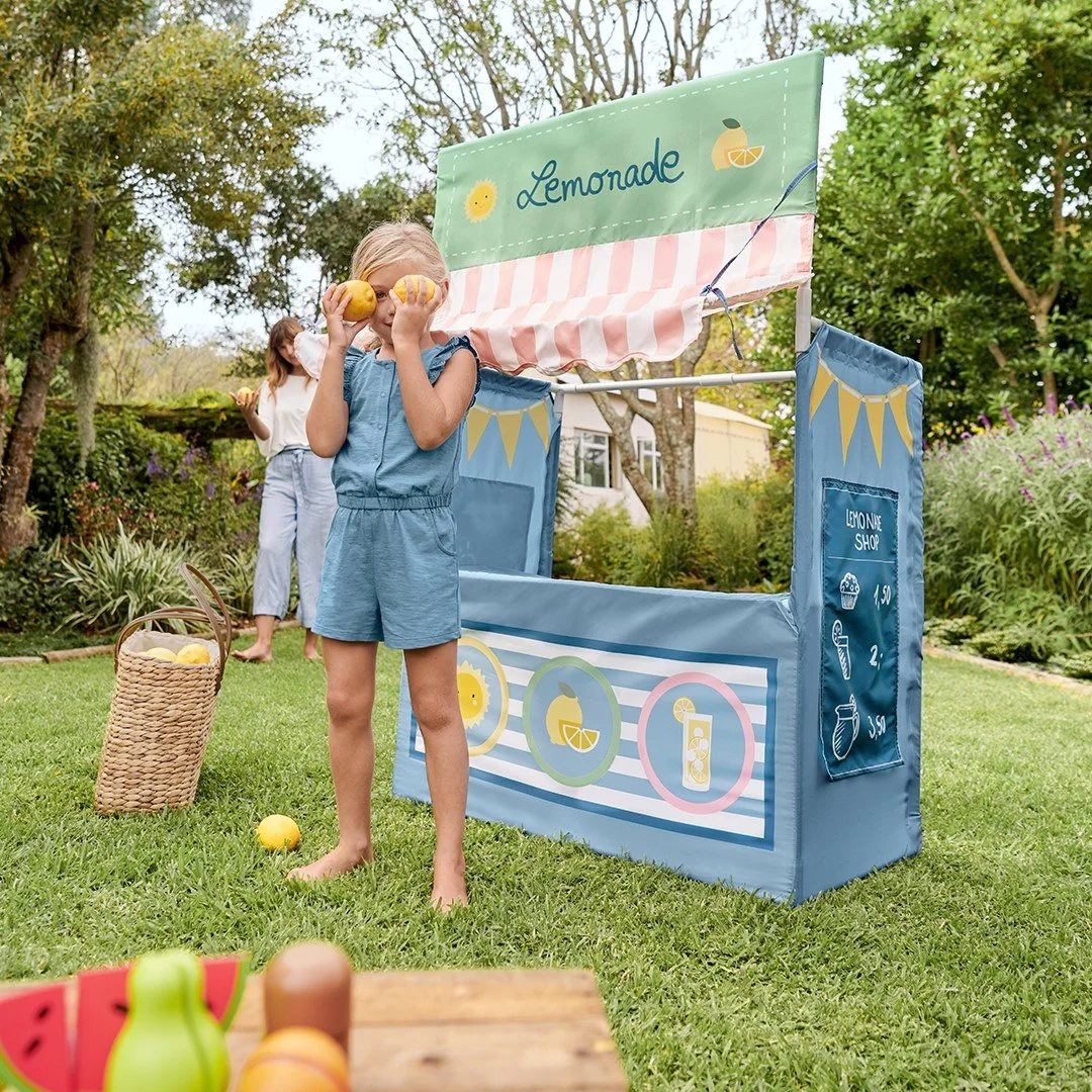 Child in blue outfit holding two lemons in front of a lemonade stand, with a woman and garden in the background.