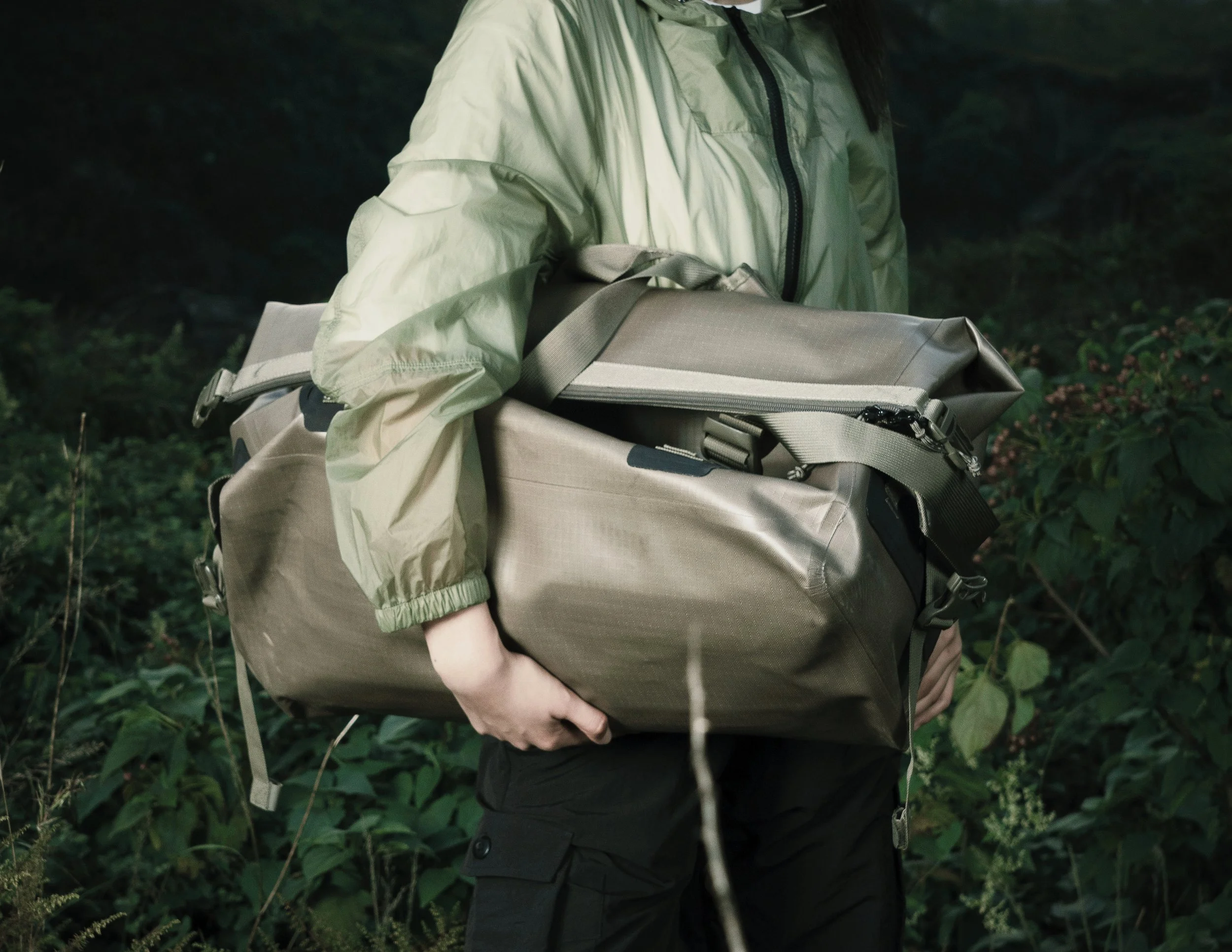 Person wearing a green jacket holding a large, gray waterproof duffel bag in a forest setting.