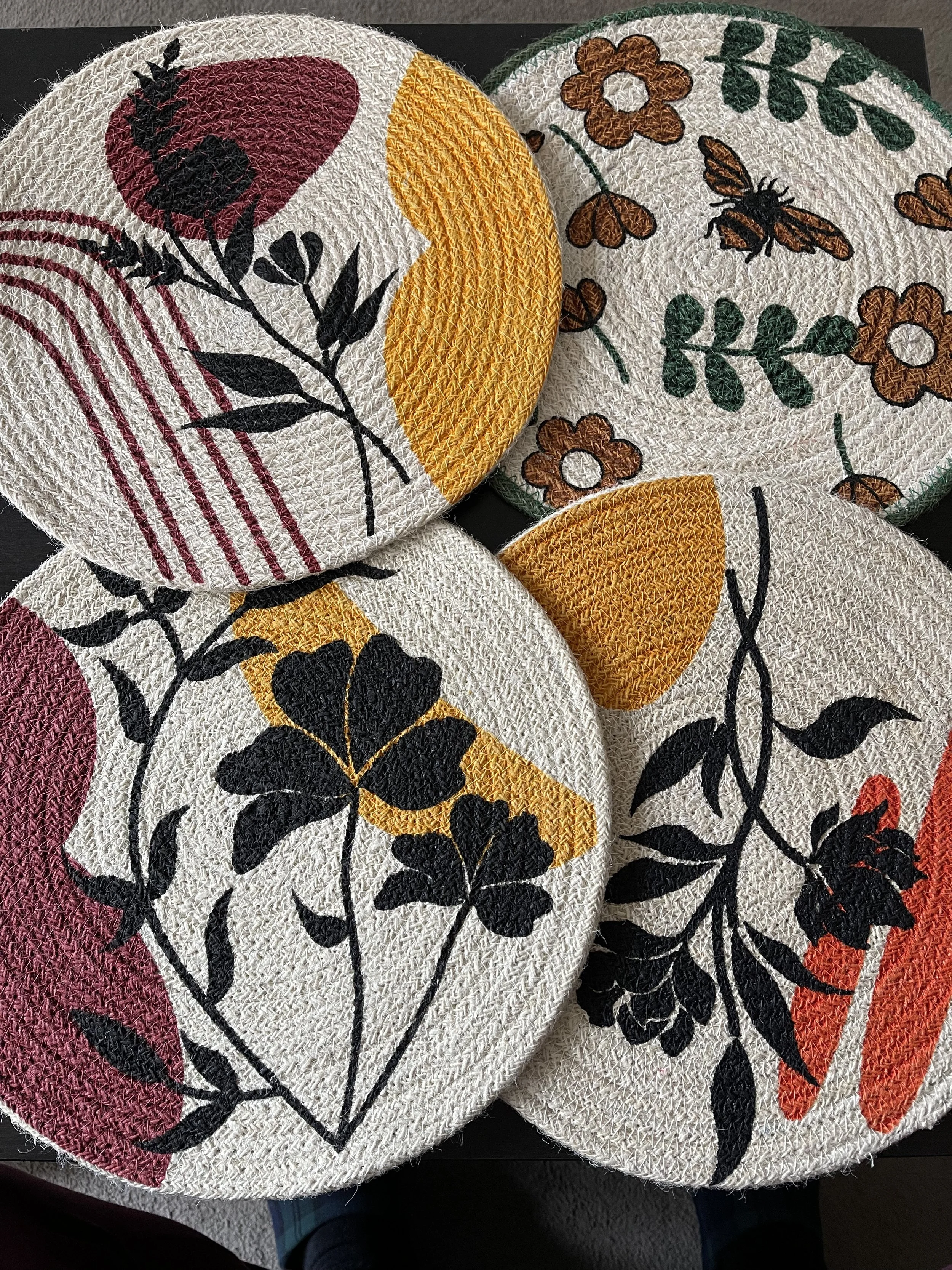 Four round fabric coasters with floral and abstract patterns in red, yellow, black, and green on a white textured background.