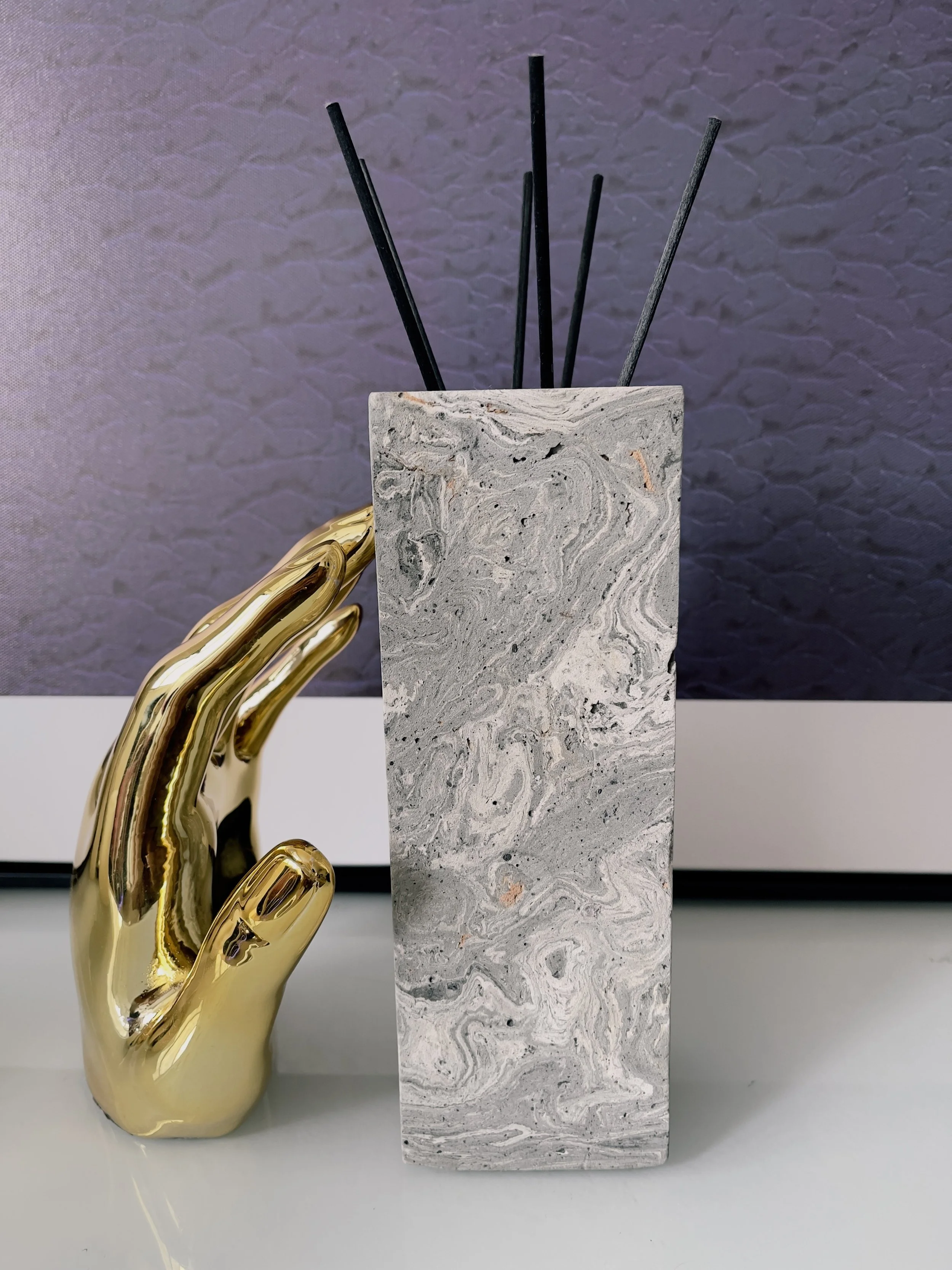 A decorative grey marble-patterned vase holding black reed sticks, with a shiny gold hand-shaped sculpture beside it, placed on a white surface against a dark textured background.