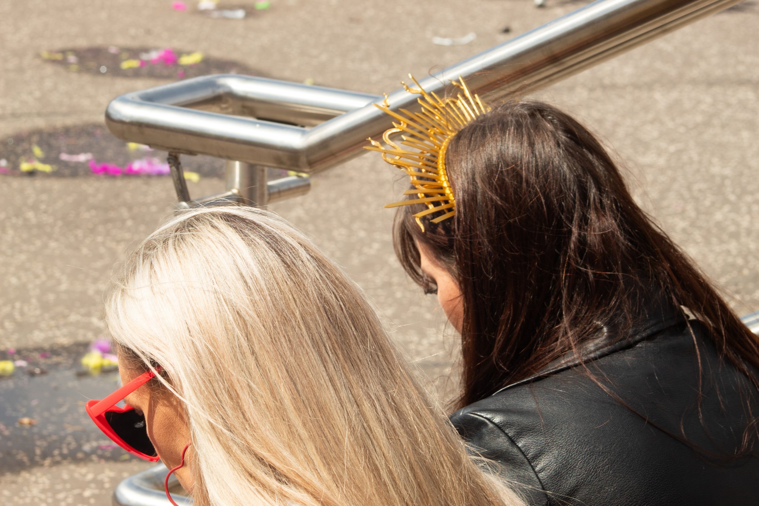 Two women with different hair colors, one with blonde and the other with brown hair, sitting closely on a bench outdoors, one wearing red glasses and the other a gold headband with radiating spikes.