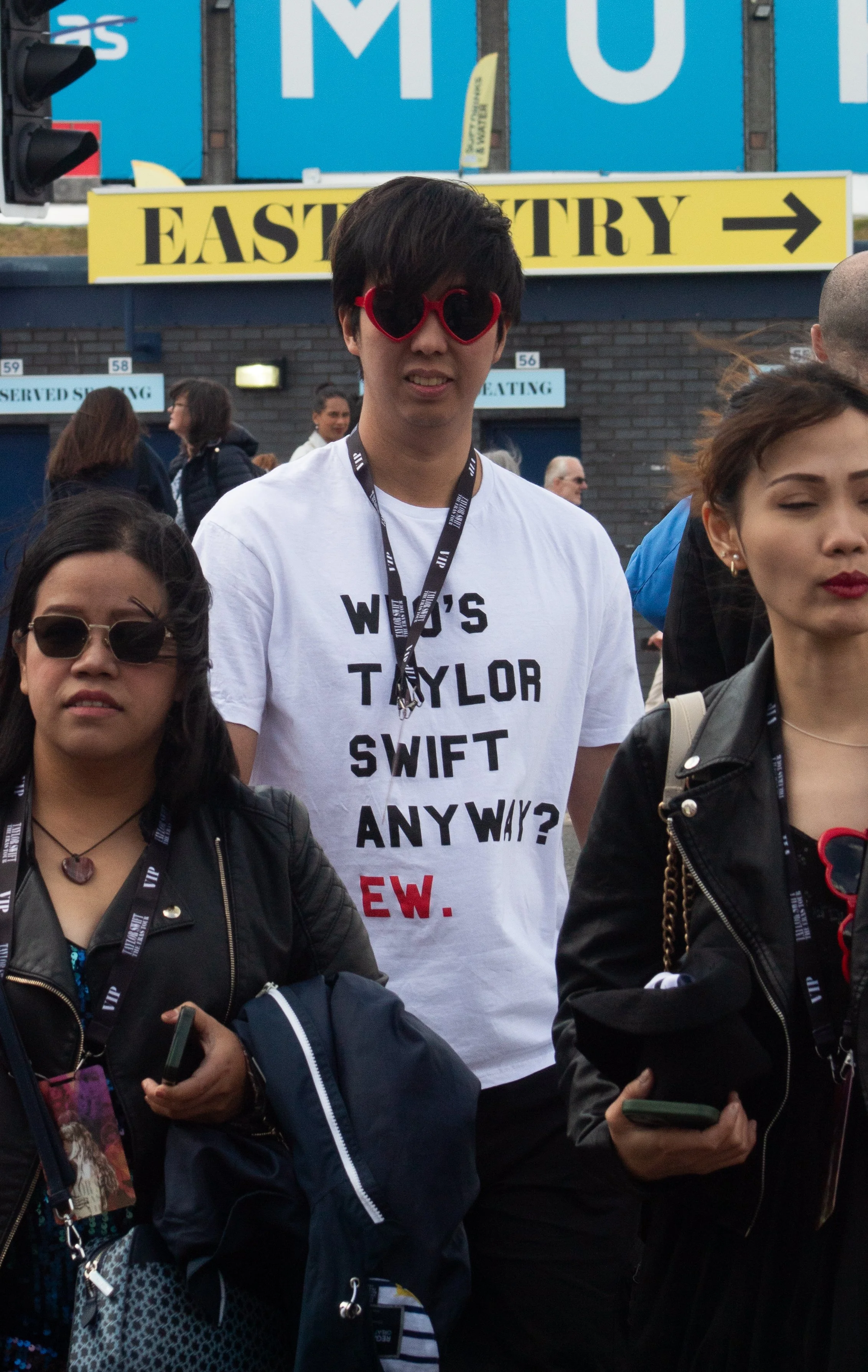 A group of people standing outdoors at an event, with a young man wearing red sunglasses and a white T-shirt that has black and red text, among others. The background features a sign that says 'EAST ENTRY' and a crowd of people.