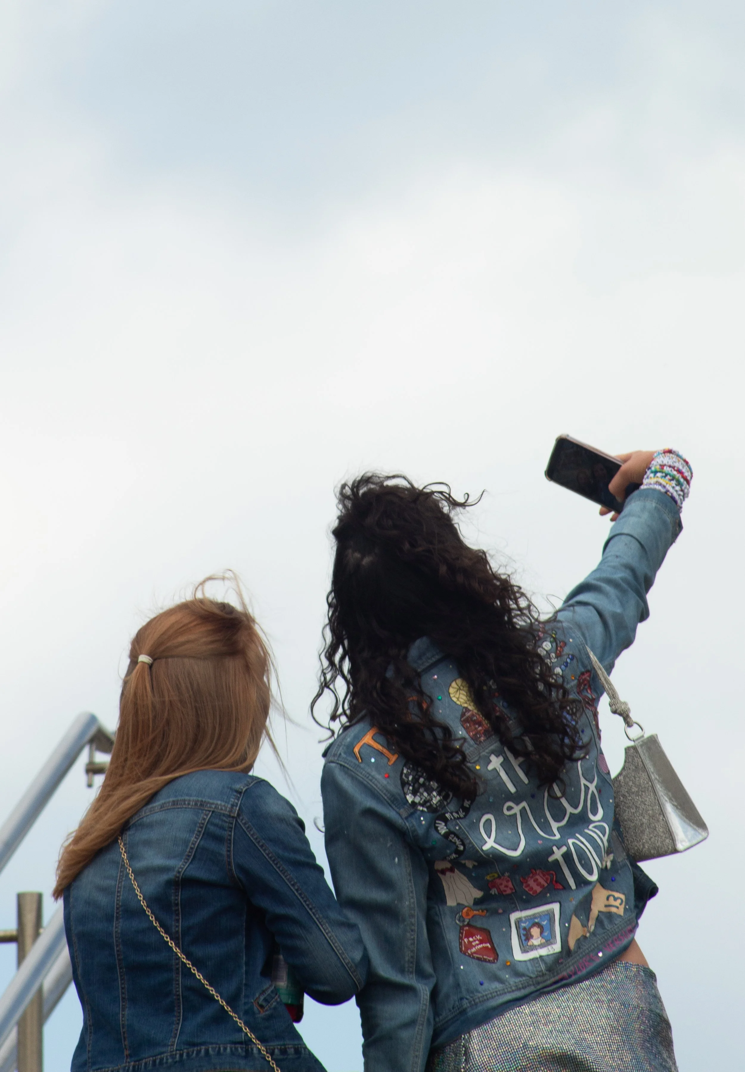 Three young women outdoors, two are taking a selfie with a smartphone while the third looks on. They are all wearing denim jackets, and one has colorful beaded bracelets and a tote bag.
