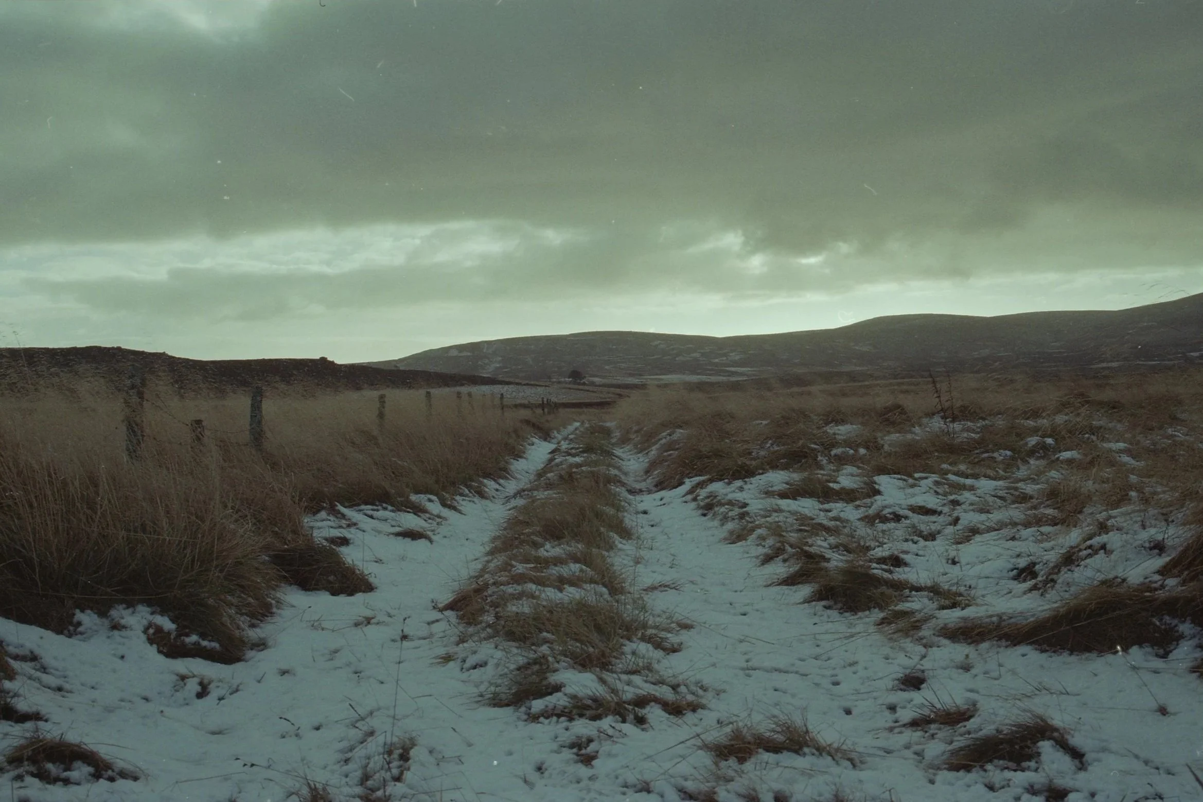 A snowy dirt path leading through a grassy hillside landscape under a cloudy sky.