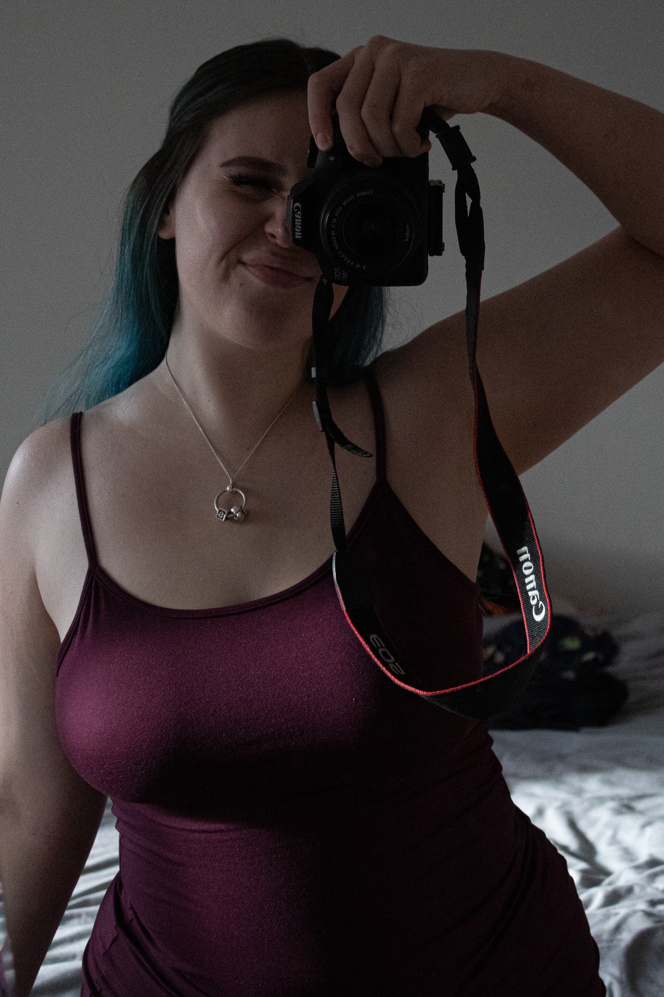 Girl with dyed hair taking a mirror selfie in a dimly lit room, smiling and wearing a purple tank top and a necklace with a key pendant.