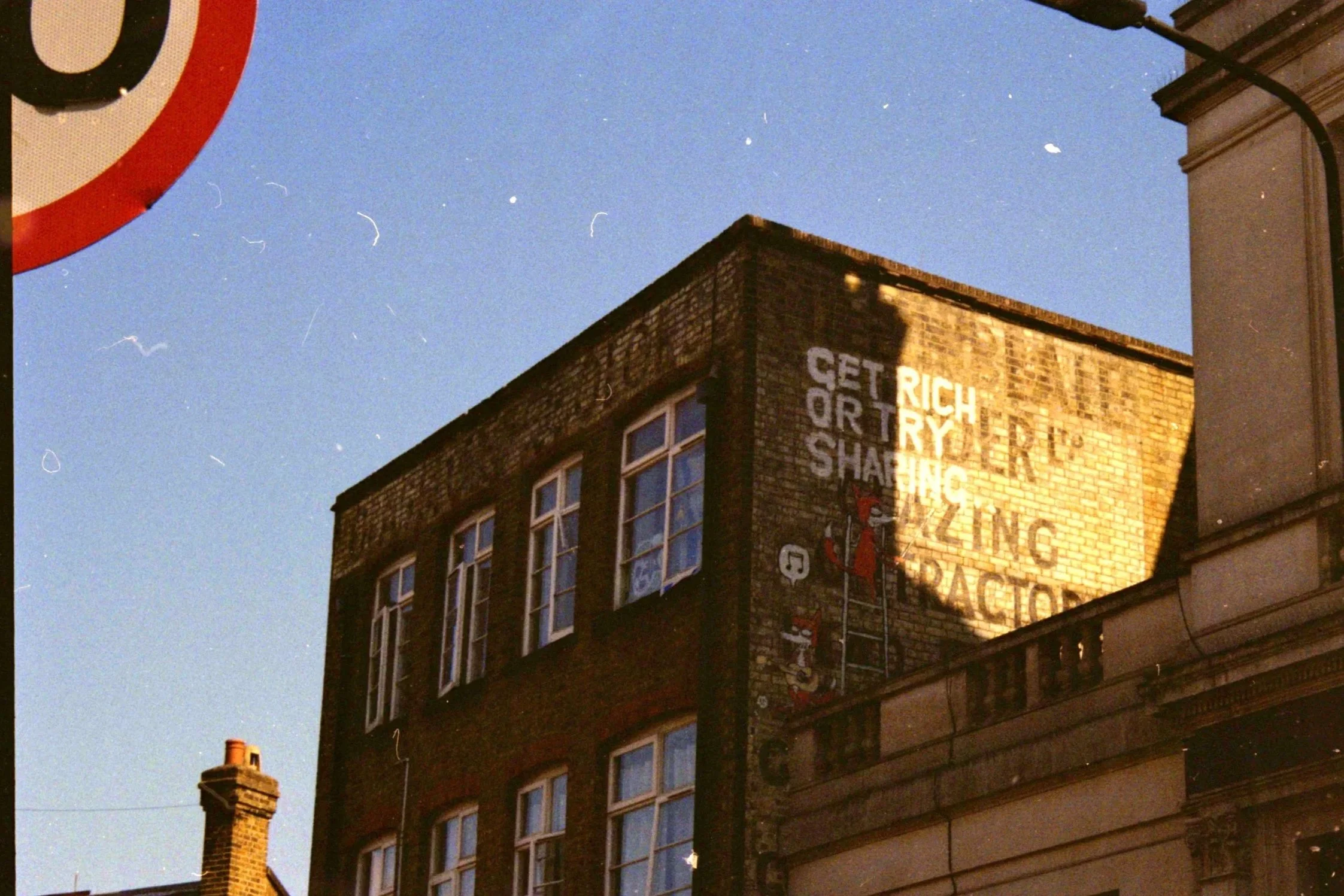 The image shows the upper part of an old brick building with large windows, some of which are open. A portion of a street sign with a red, white, and black design is visible at the top left corner, and a chimney is seen on the left side of the roof. 