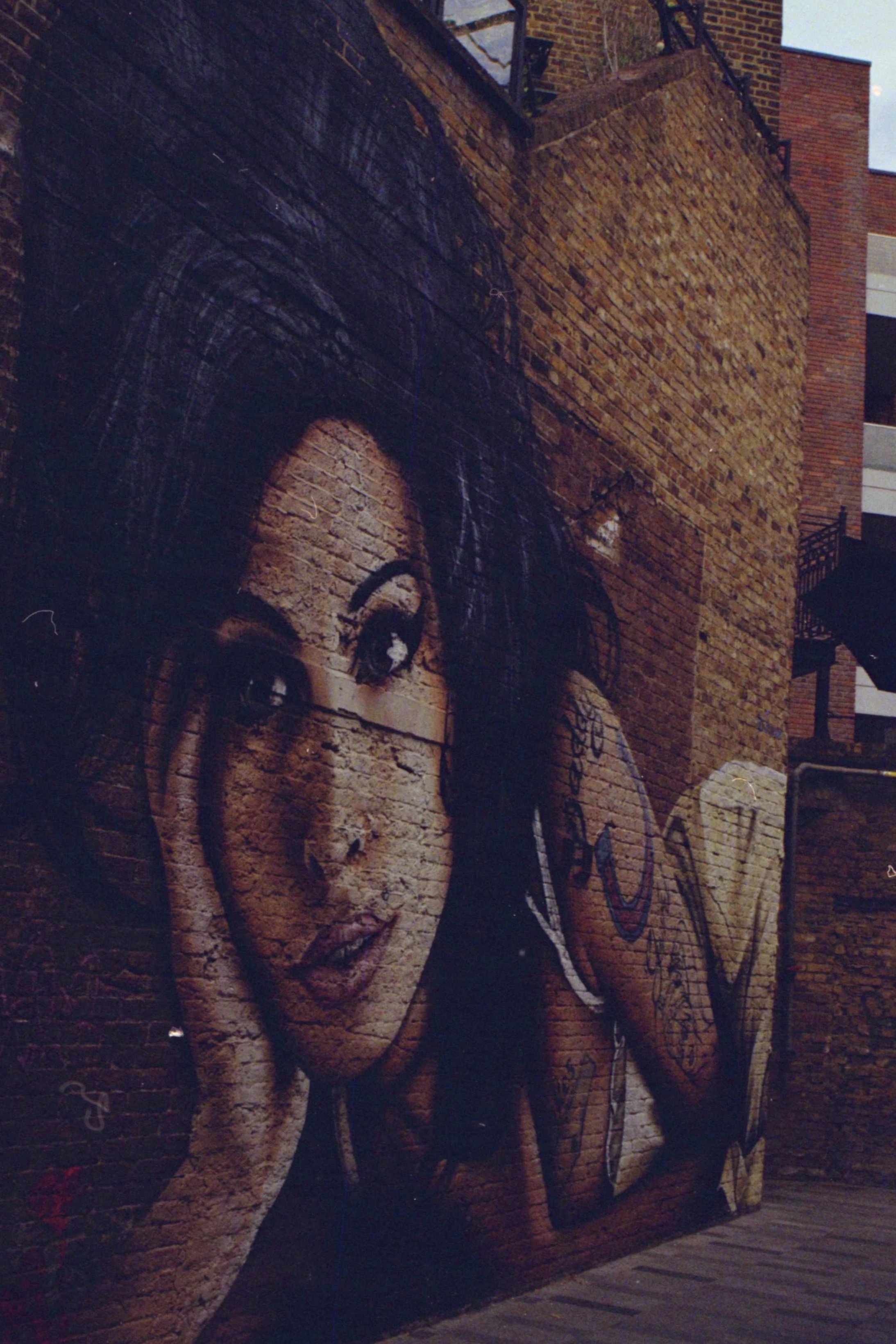 Street art mural of a woman's face painted on a brick wall, with graffiti tags on the side.