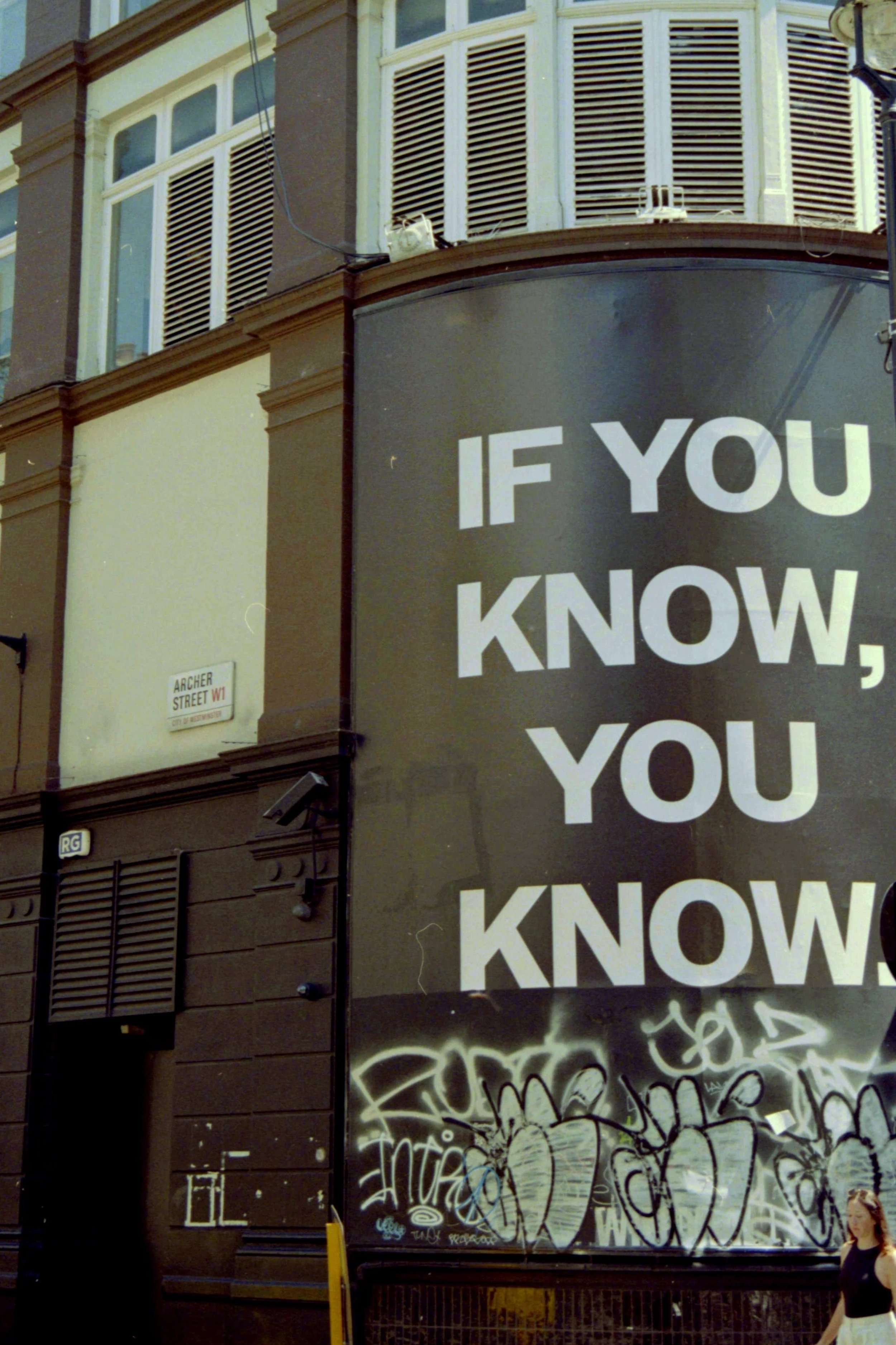A city street corner with a large black billboard that has white text reading 'IF YOU KNOW,' a building with bay windows and window shutters, a street sign that reads 'ARCHER STREET W1,' a graffiti-covered wall, and a woman walking on the sidewalk in