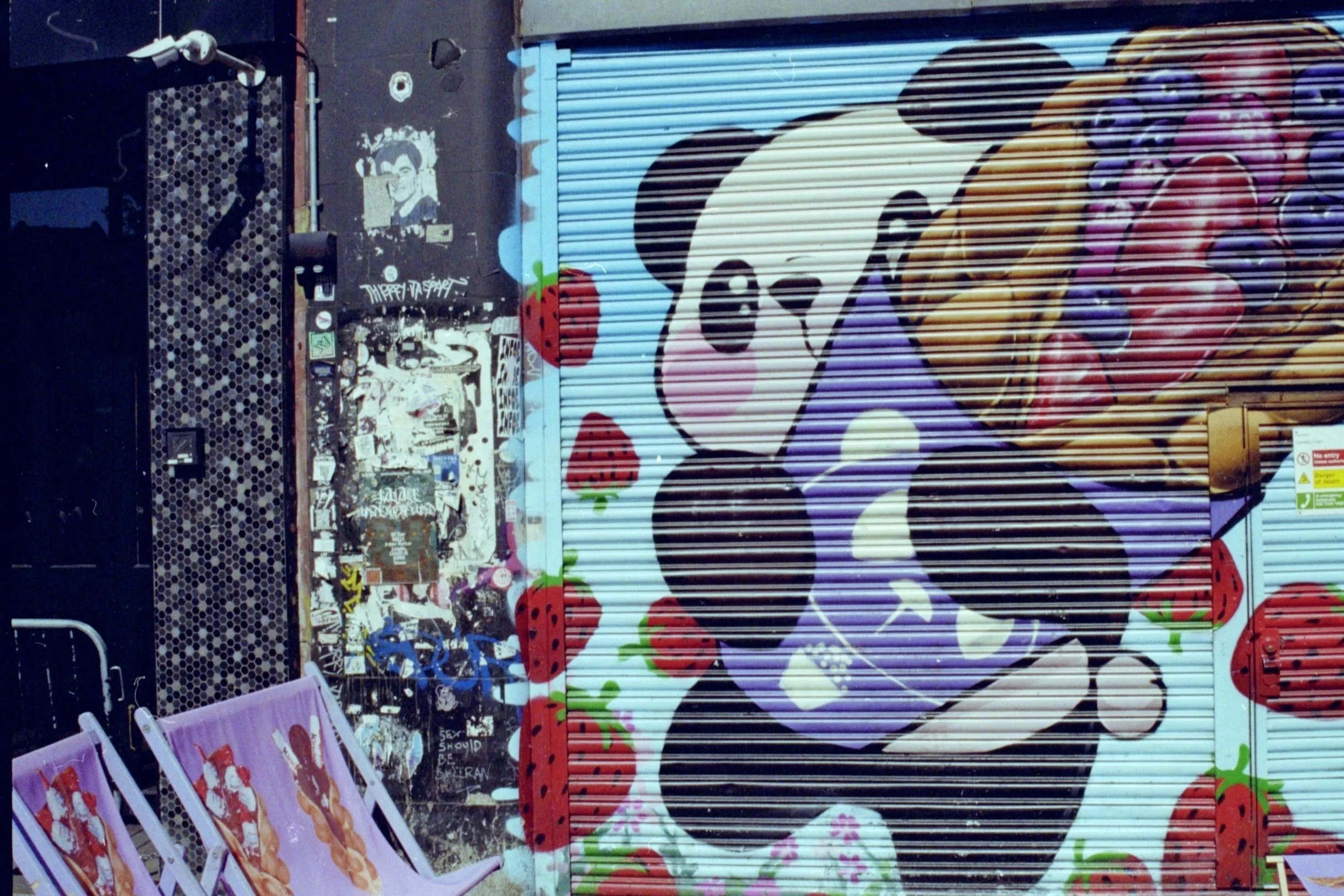 Street art mural of Mickey Mouse with strawberries, painted on a metal shutter, with various posters and graffiti surrounding it.