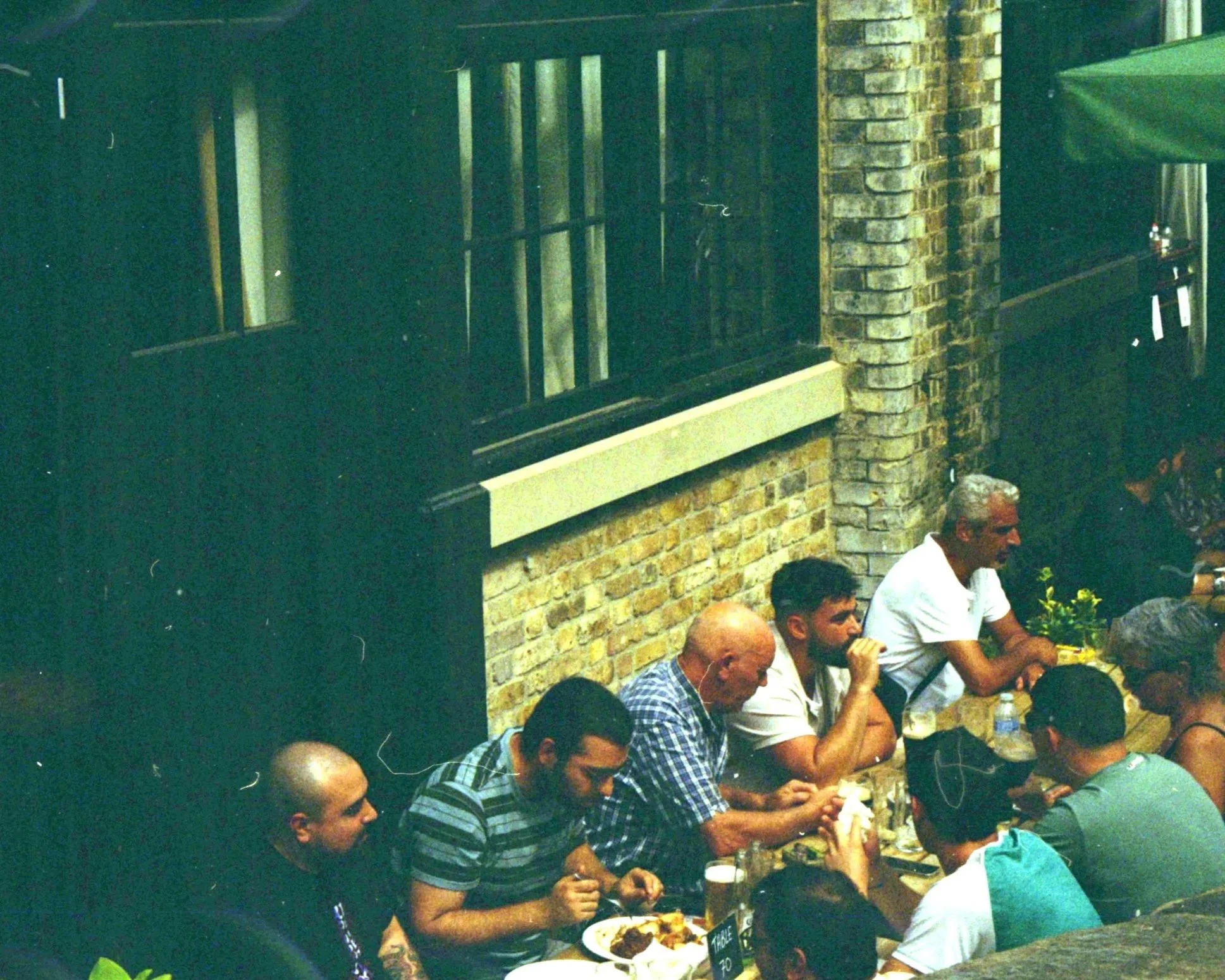 People sitting at a long table outdoors, having a meal, with brick walls and green umbrellas in the background.