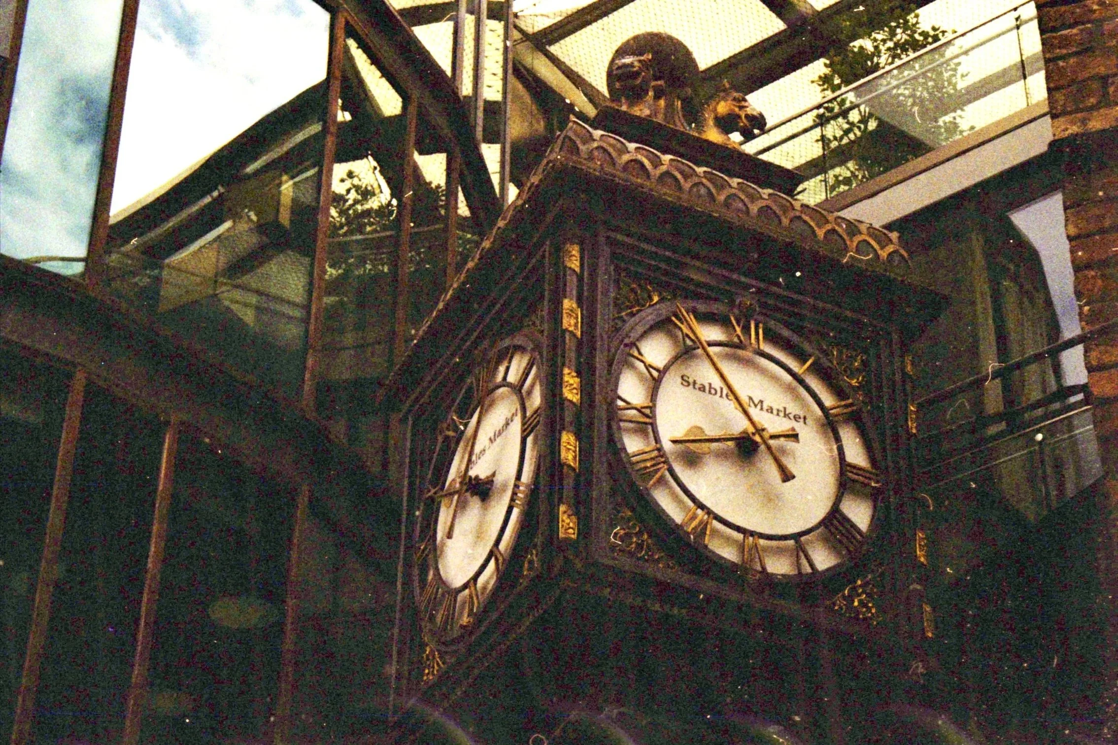 A decorative clock with Roman numerals and gold accents, labeled "Stone Market," surrounded by ornate details and greenery, with reflections showing trees and sky.