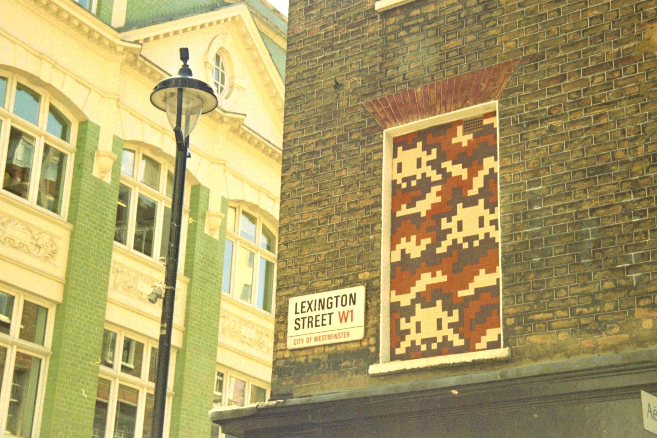 Street scene featuring a decorative brick wall with a pixelated art piece, a street sign for Lexington Street, and a city of Westminster label. A vintage street lamp is visible in front of an ornate building with large windows and green tiled exterio